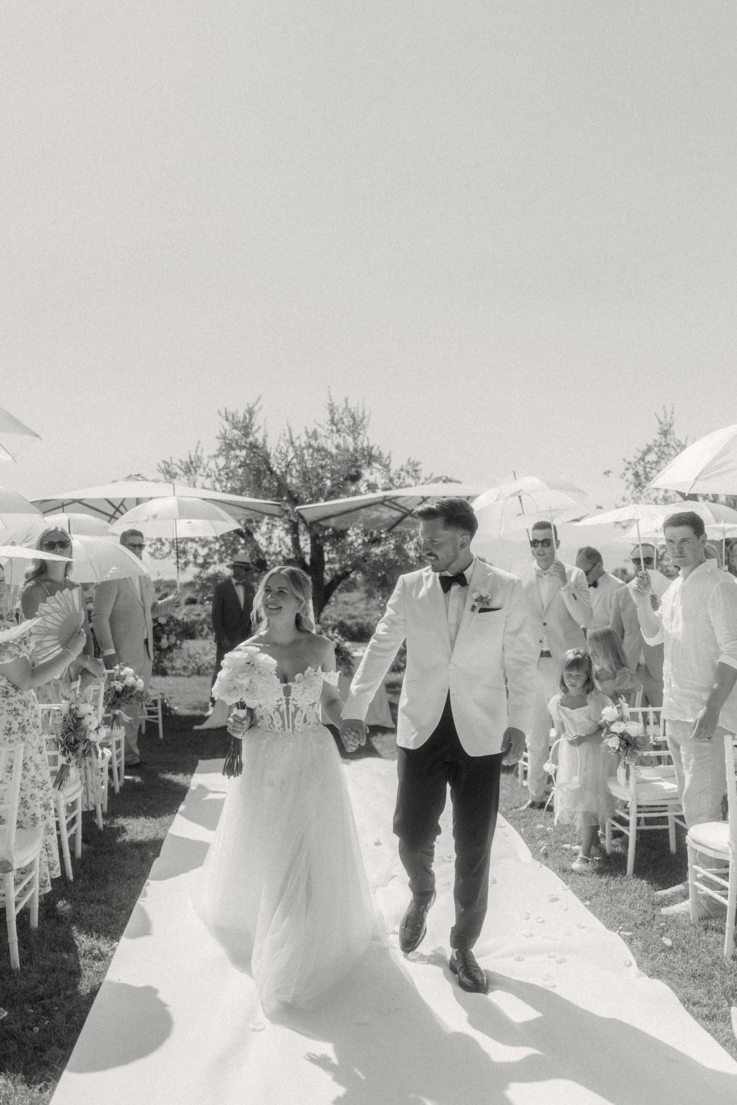 Black and white photo of bride and groom walking down outdoor wedding aisle with guests holding parasols.