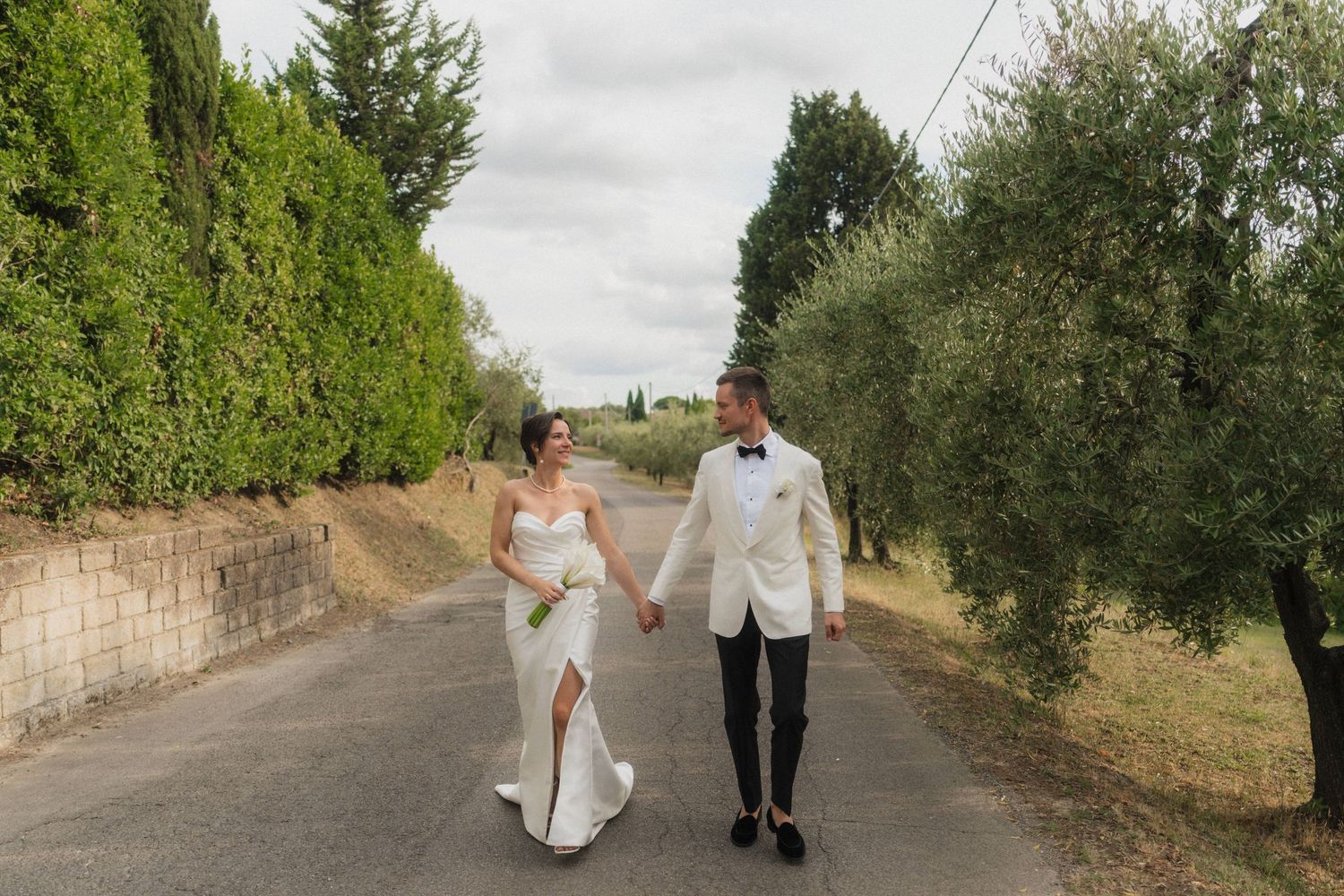Bride and groom walking hand in hand down rural road lined with olive trees.