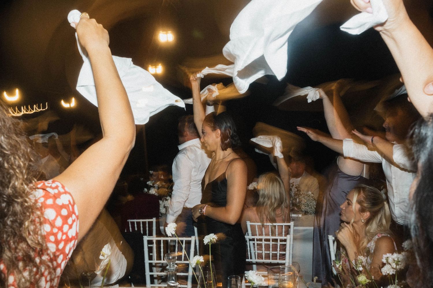 Excited wedding guests wave white napkins during lively indoor reception party at night.
