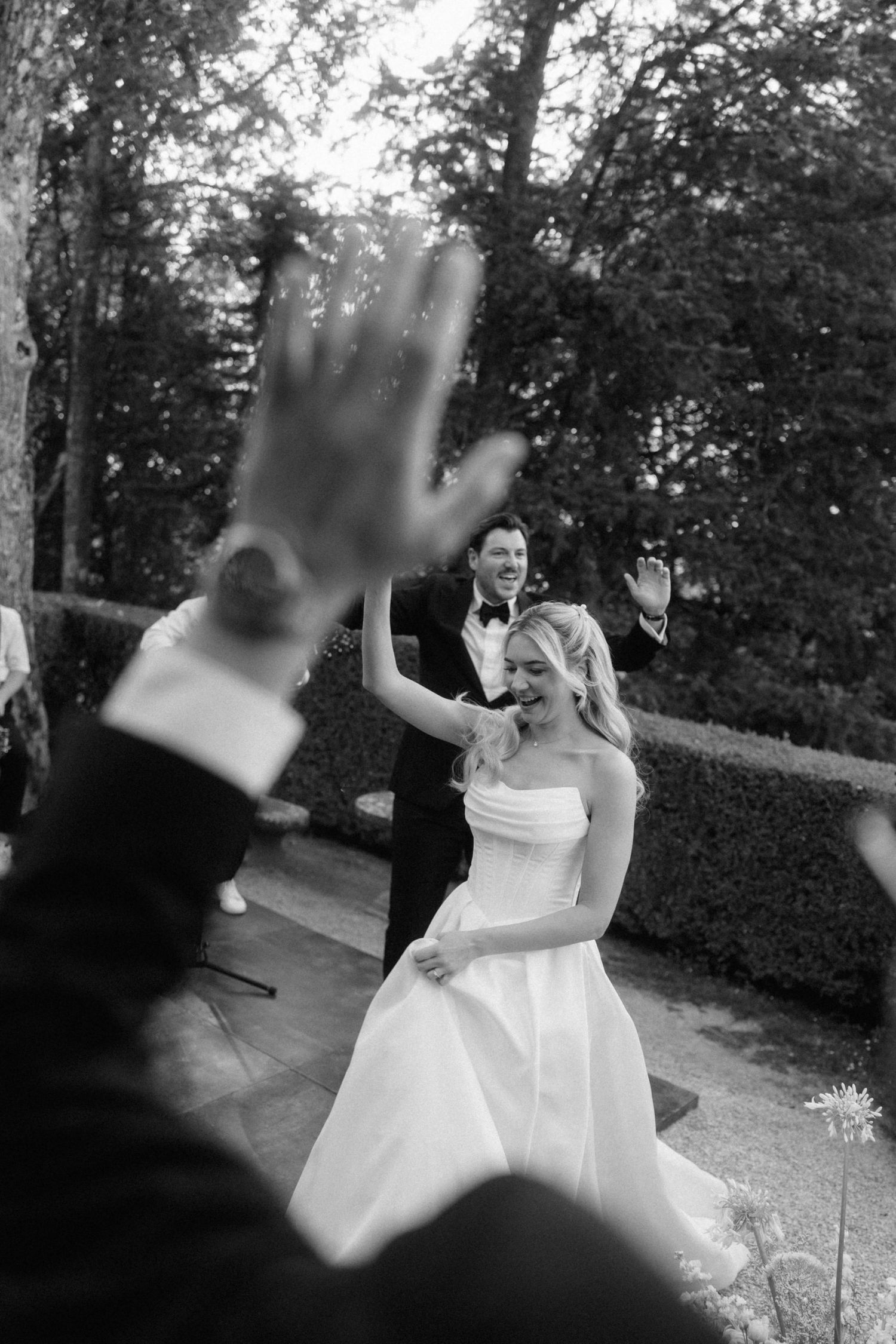 Black and white photo of smiling bride in strapless gown celebrating with guests at outdoor wedding.