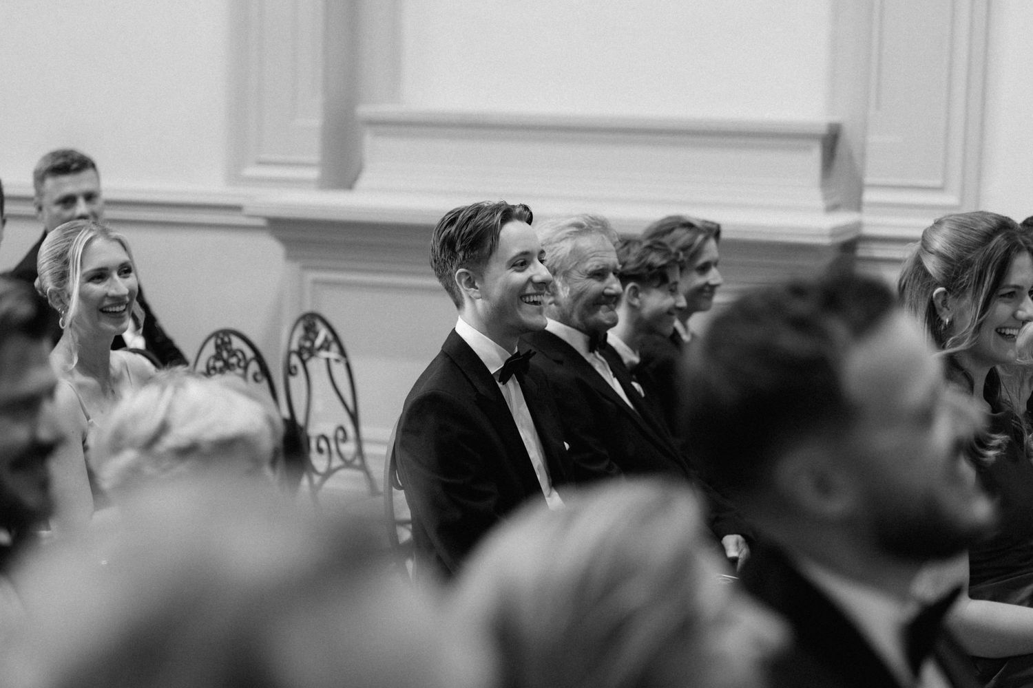 Wedding guests smiling and laughing during ceremony in black and white photo at elegant venue.