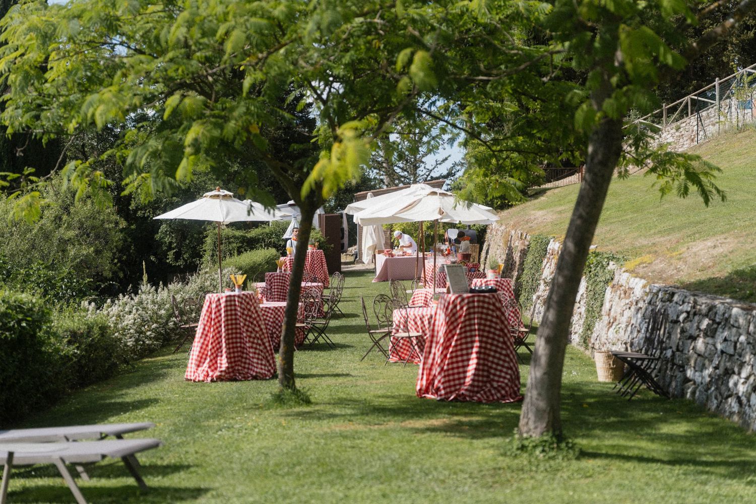 Garden wedding reception setup with red gingham tablecloths and white umbrellas on a sunny green lawn.