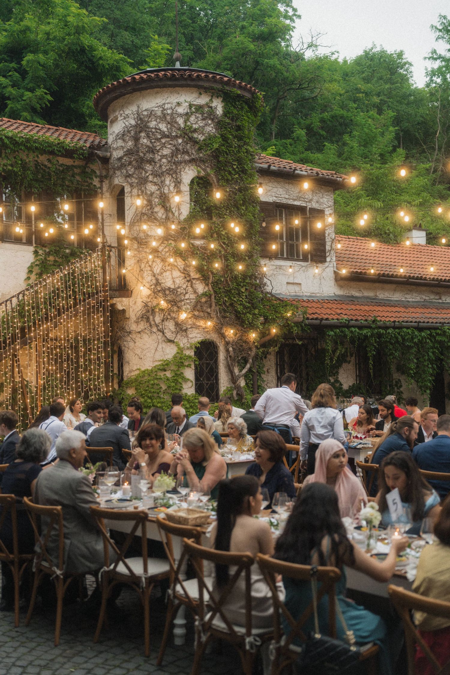 Romantic outdoor wedding reception dinner under string lights at ivy-covered villa at dusk.