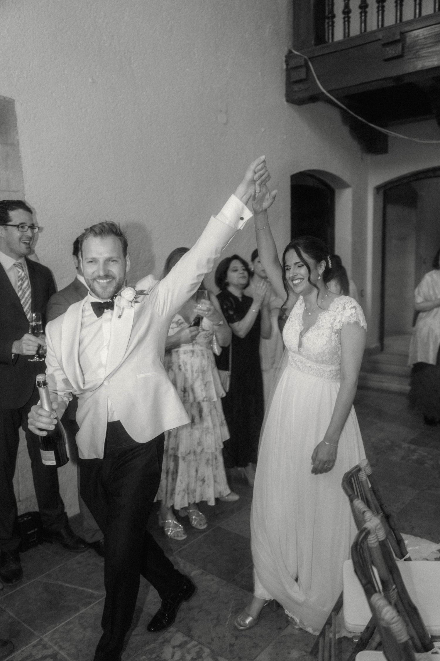 Black and white photo of a groom raising his hand in celebration with bride and guests at a wedding reception.