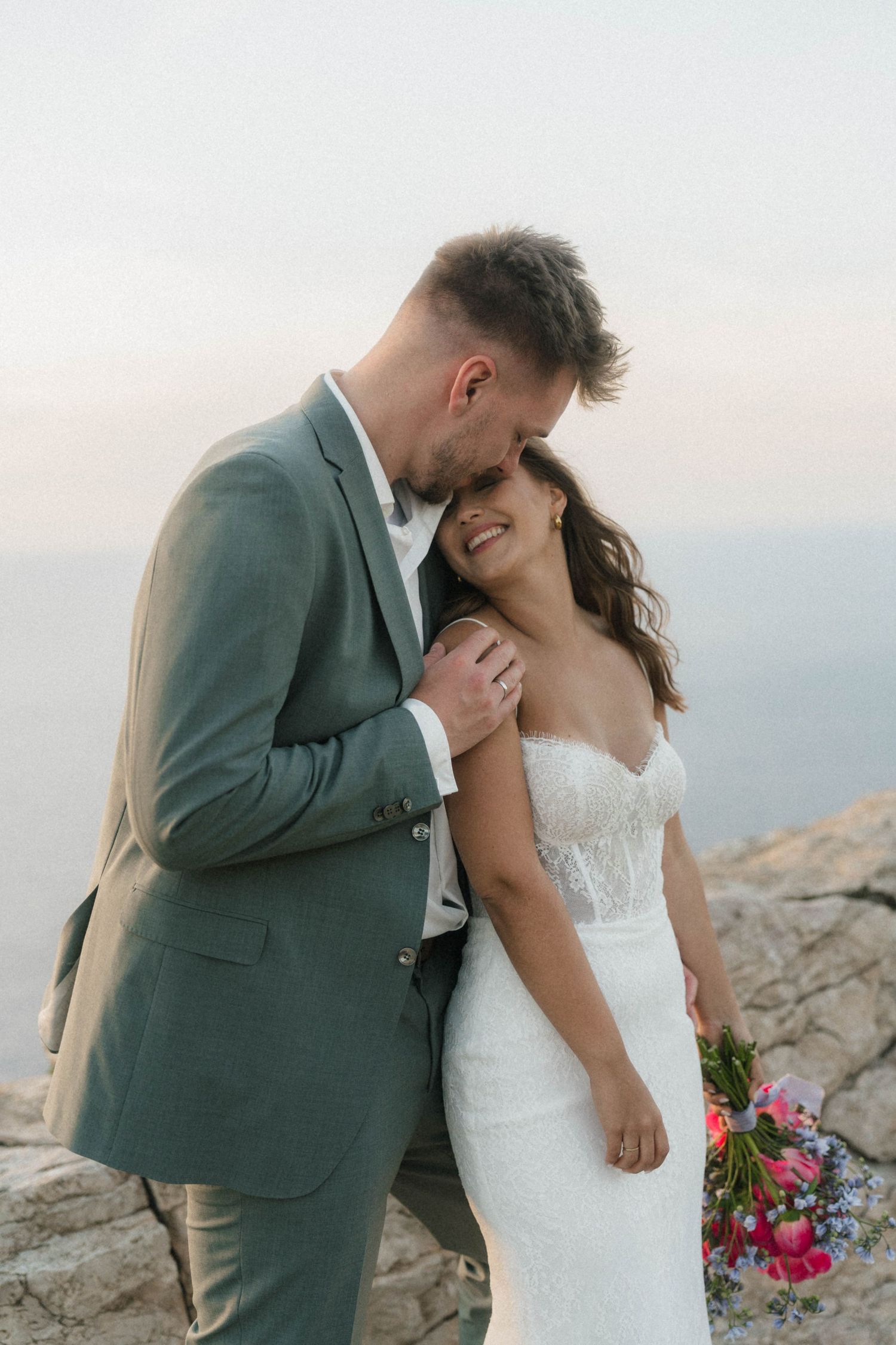 Bride and groom share a tender moment on rocky cliffs, groom in gray suit kissing brides head in white lace gown.