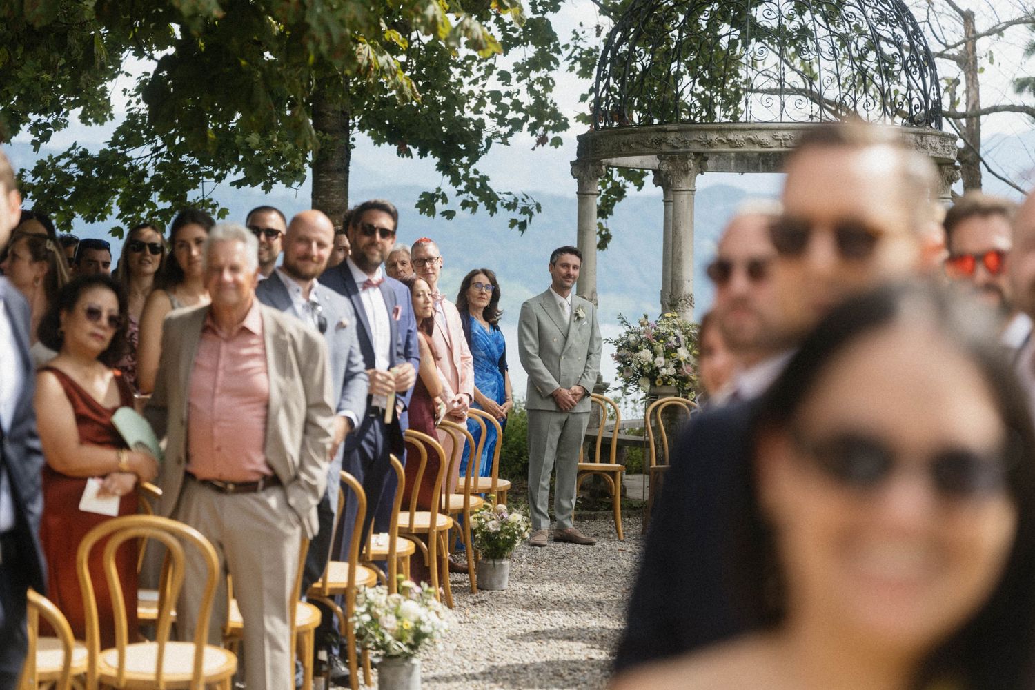 Wedding guests seated in golden chairs along an outdoor aisle with a scenic lake view in the background.