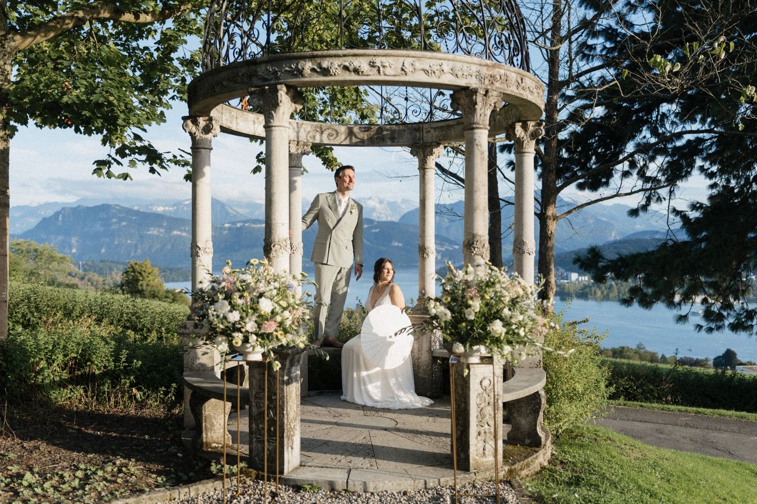 Bride and groom stand under elegant stone gazebo with white floral arrangements, overlooking a scenic lake and mountains.