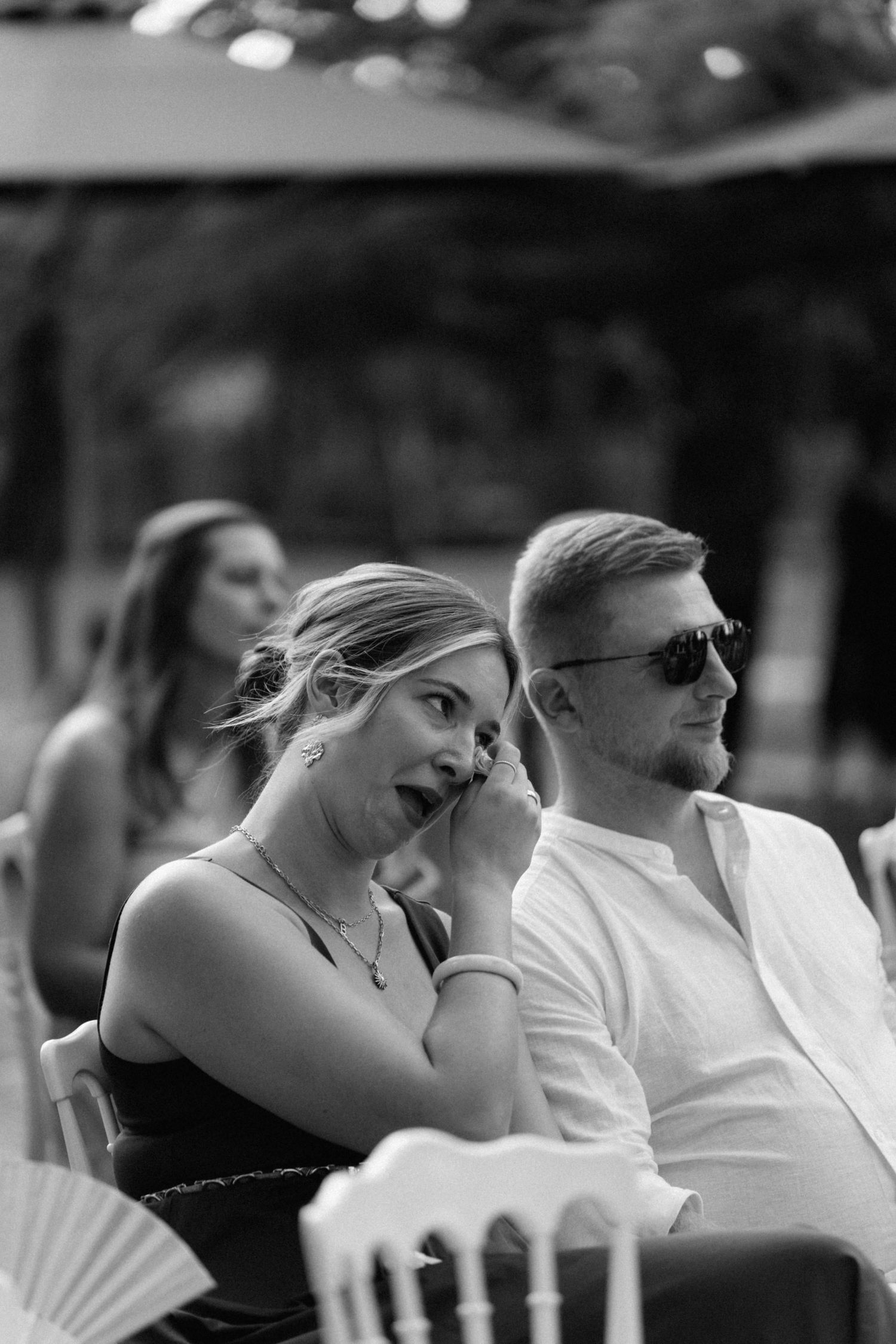 Black and white photo of surprised woman wiping eye beside man in sunglasses at outdoor wedding ceremony.