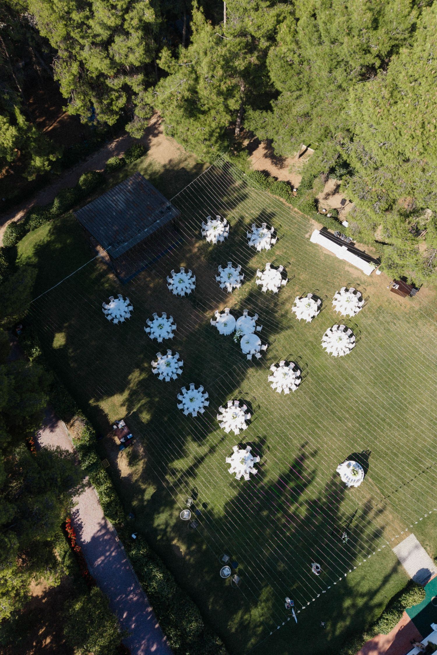 Aerial drone view of white round reception tables arranged on a green lawn surrounded by tall pine trees at an outdoor wedding.