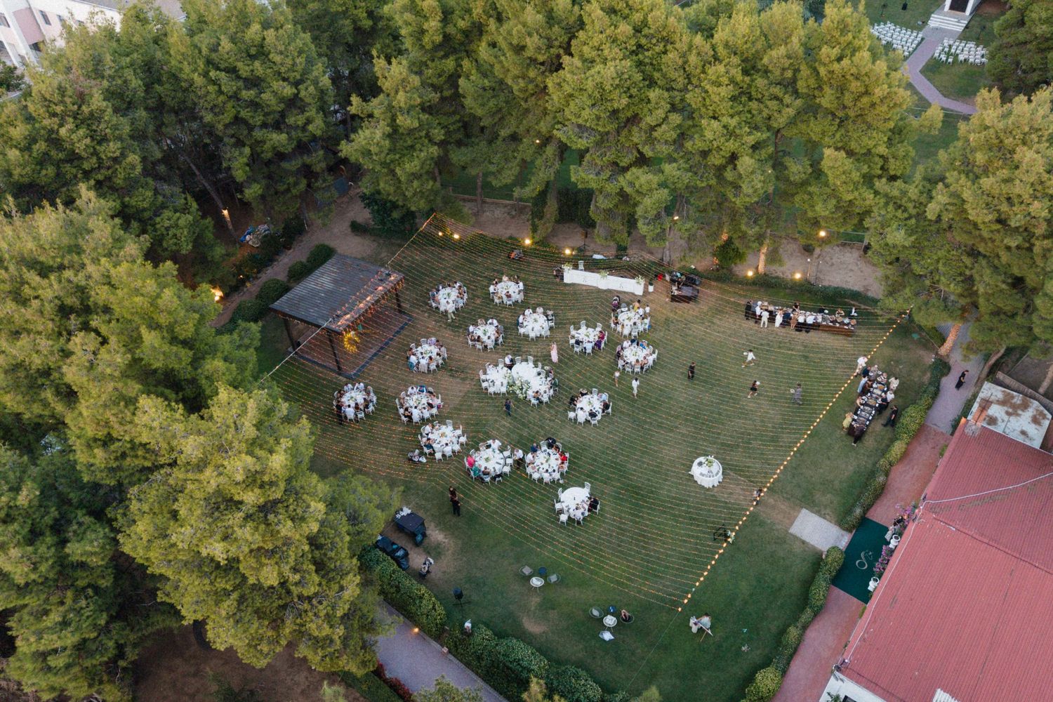 Aerial view of an elegant outdoor wedding reception with white round tables on green lawn surrounded by tall trees.