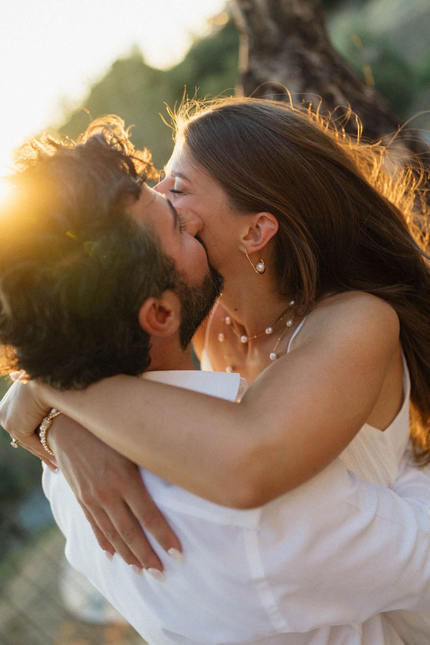 Couple sharing a romantic kiss at golden hour sunset, woman wearing white dress and pearl jewelry embracing man outdoors.
