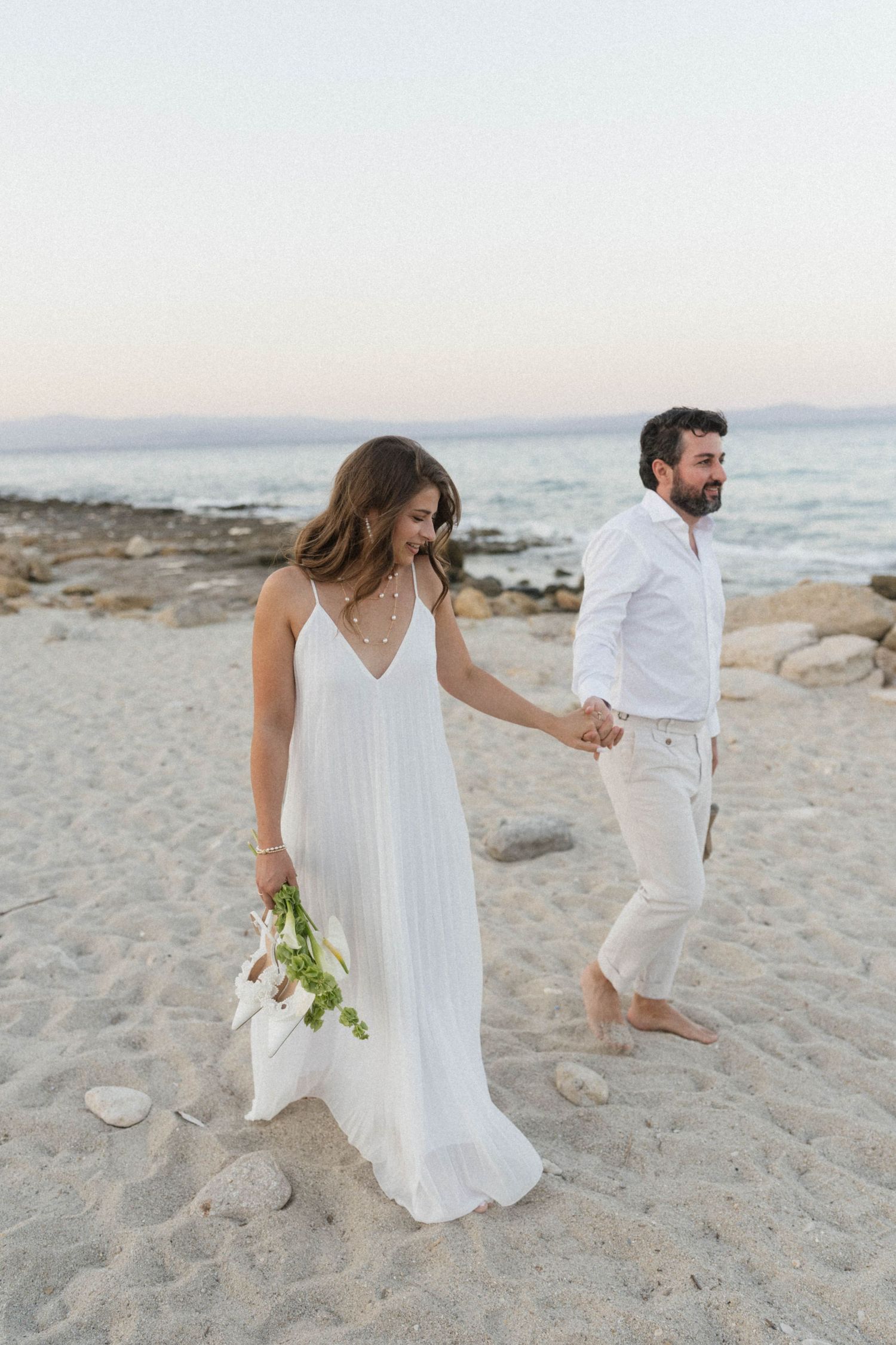 Bride in white spaghetti strap gown holding bouquet walks hand in hand with groom on sandy beach at dusk.