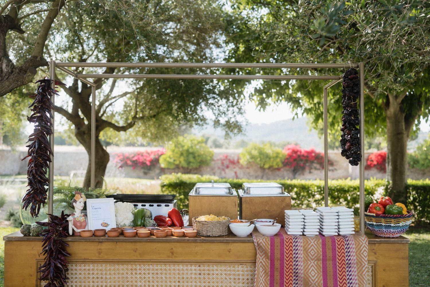 Outdoor buffet station with colorful linens, stacked white bowls, chili pepper garlands, and fresh produce under a rustic wooden frame.