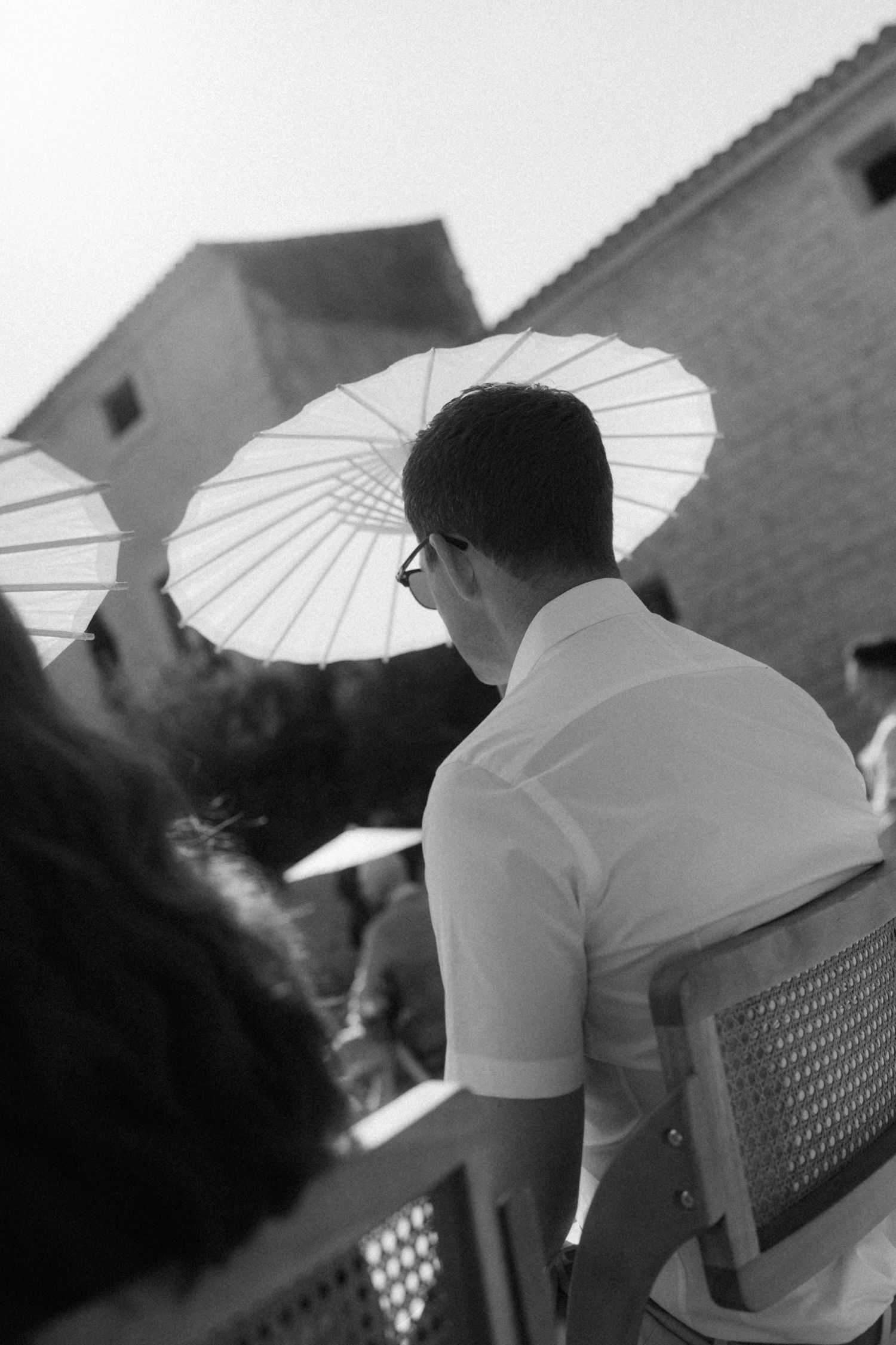 Black and white photo of wedding guest holding white parasol umbrella seated at outdoor ceremony.
