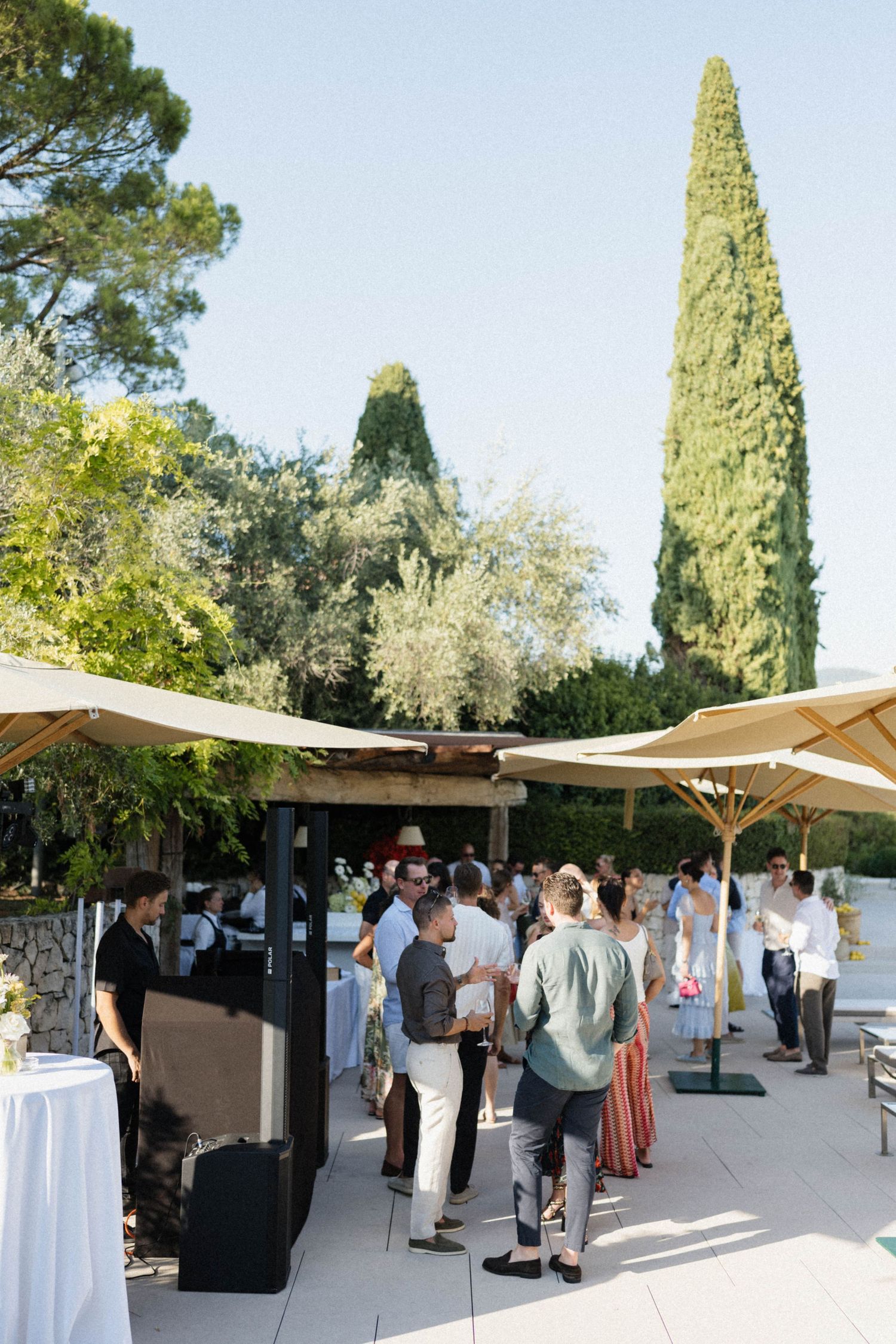 Elegant outdoor garden party with guests mingling under shade sails, surrounded by cypress trees and lush greenery.
