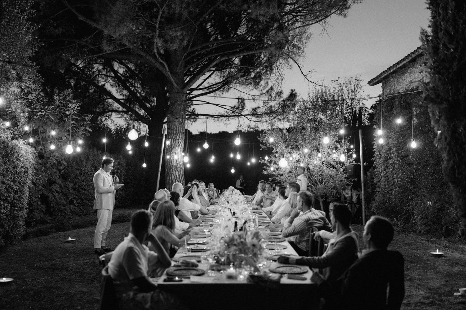 Black and white outdoor dinner party under string lights with guests seated at long table in garden at dusk.