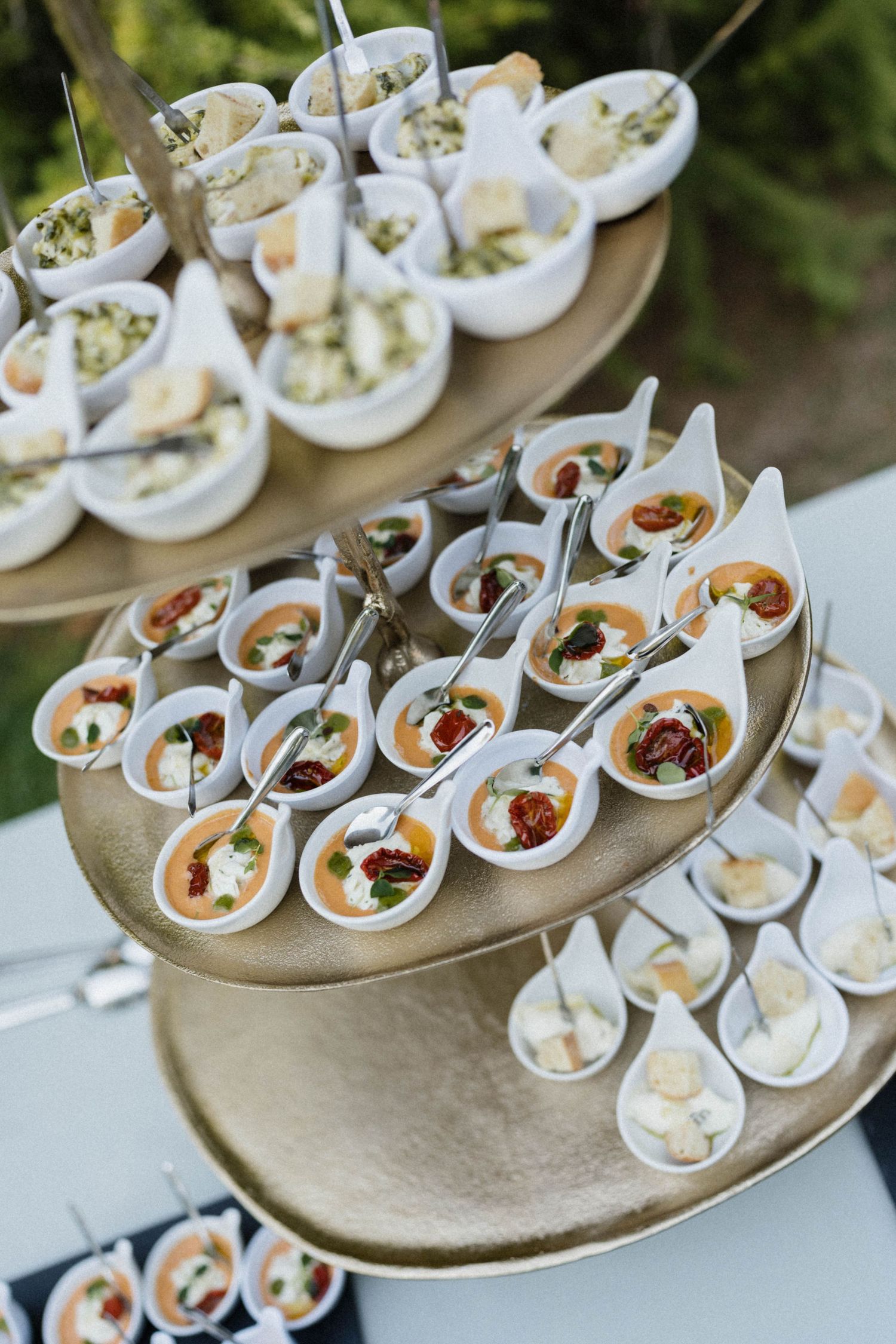Elegant three-tiered catering display with white appetizer spoons and bowls filled with gourmet bite-sized hors d\u0027oeuvres.