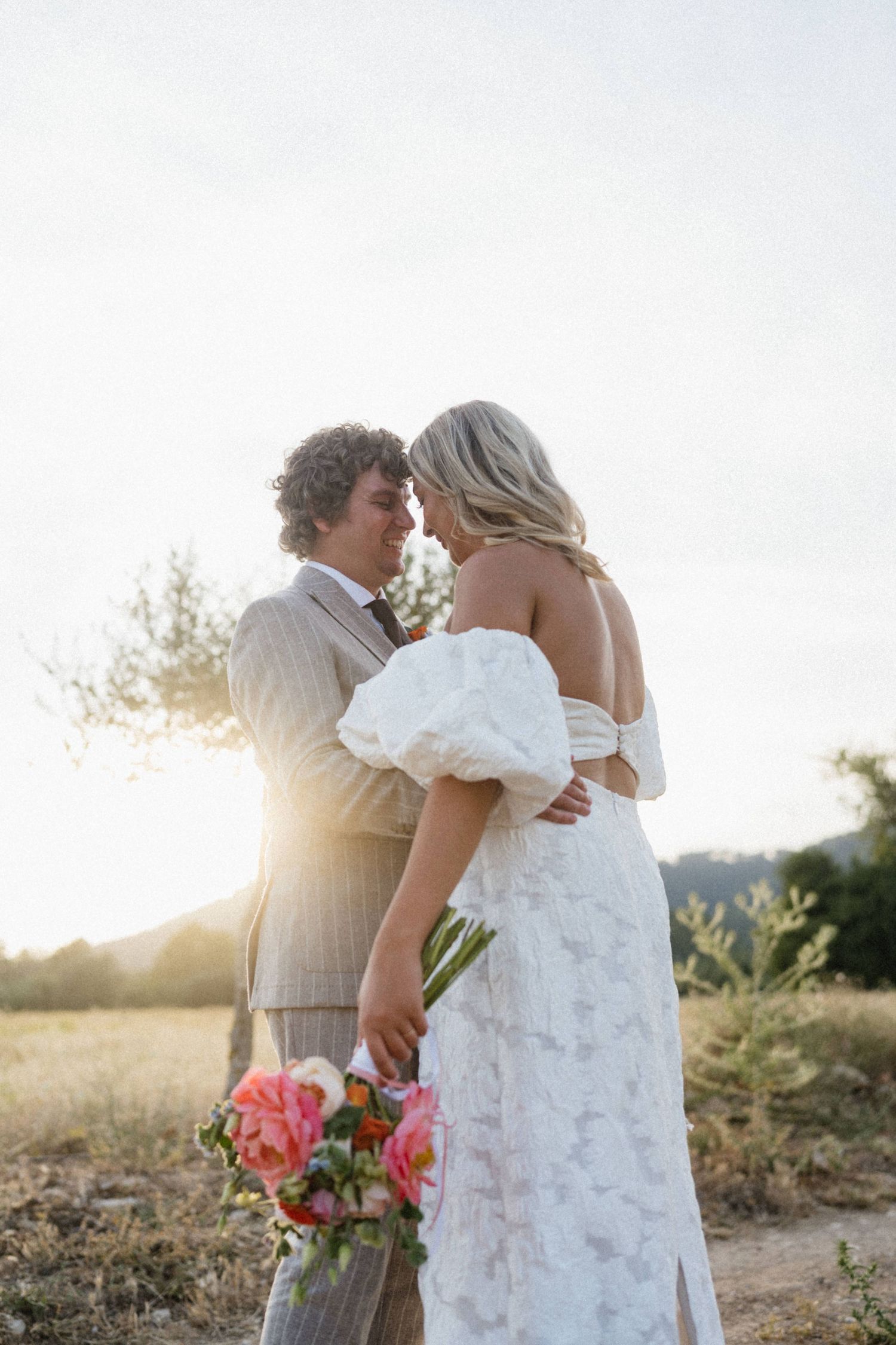Bride and groom share a romantic sunset moment outdoors, bride in off-shoulder floral gown holding pink bouquet.