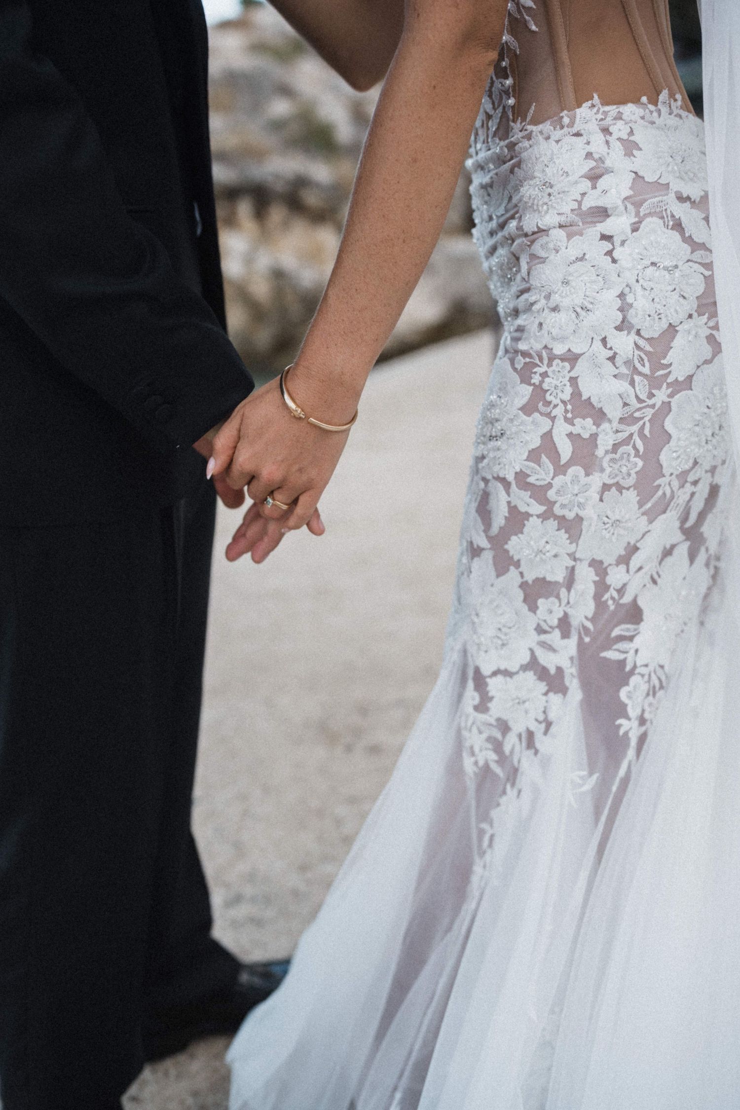 Close-up of bride and groom holding hands showing wedding ring and gold bracelet.