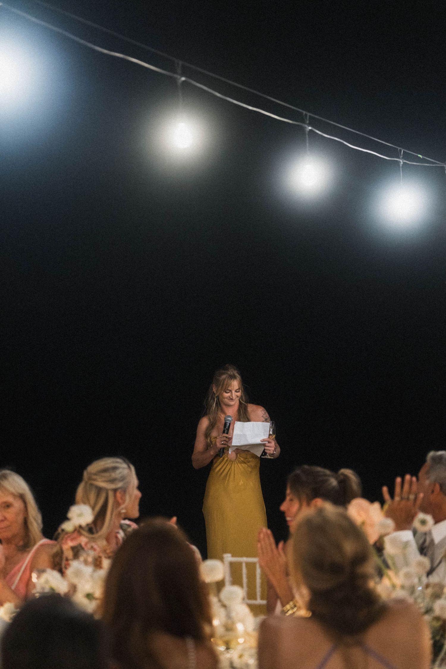 Woman in a gold dress giving a heartfelt speech at an outdoor wedding reception under string lights at night.
