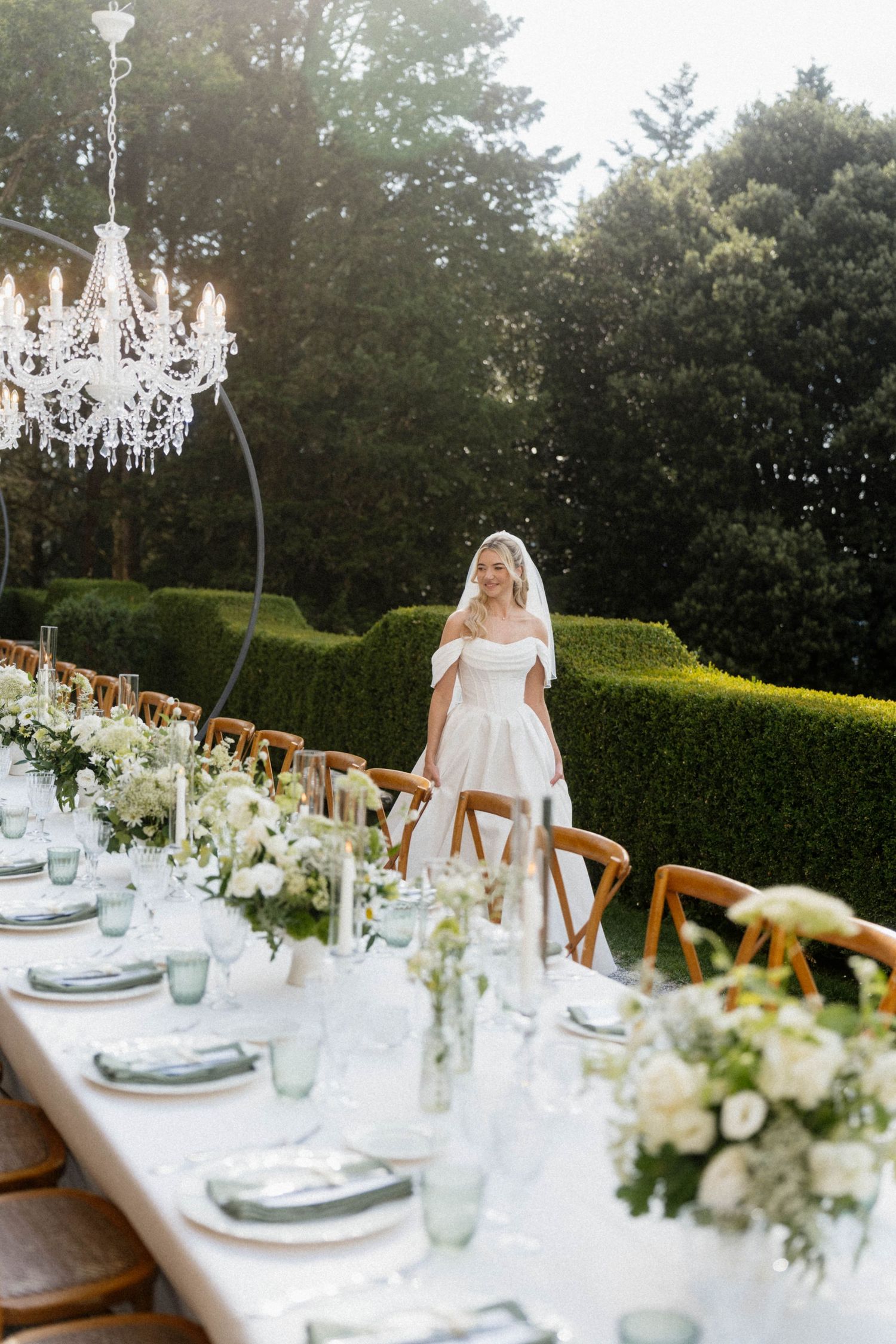 Bride in white gown standing beside elegantly set outdoor wedding reception table with crystal chandelier backdrop.