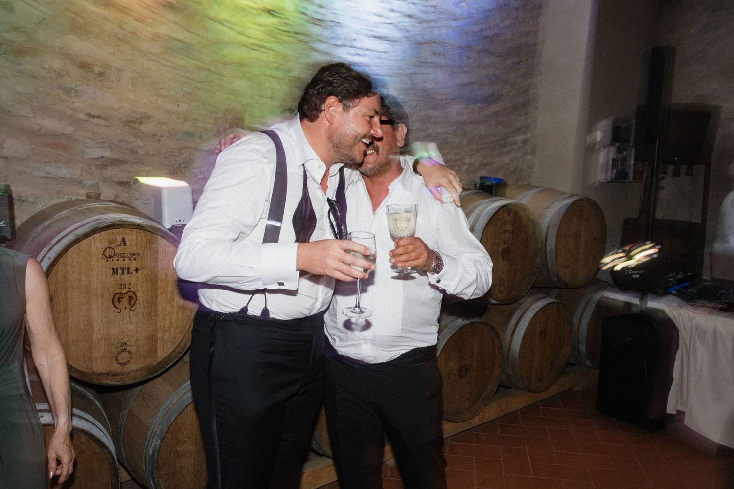 Two men in suspenders laughing and holding champagne glasses near wine barrels at wedding reception.