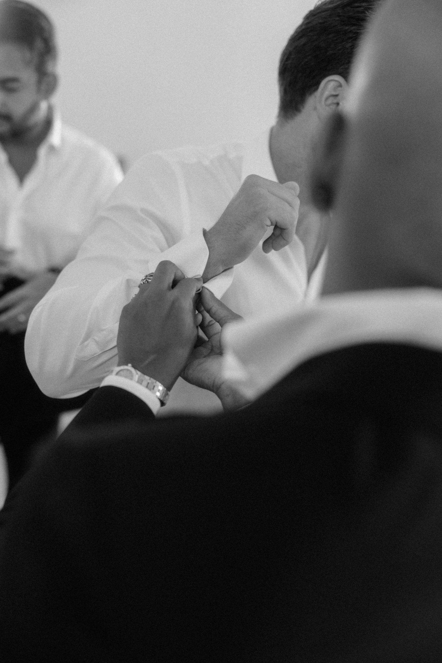 Black and white photo of groomsmen helping groom button his white dress shirt before wedding ceremony.
