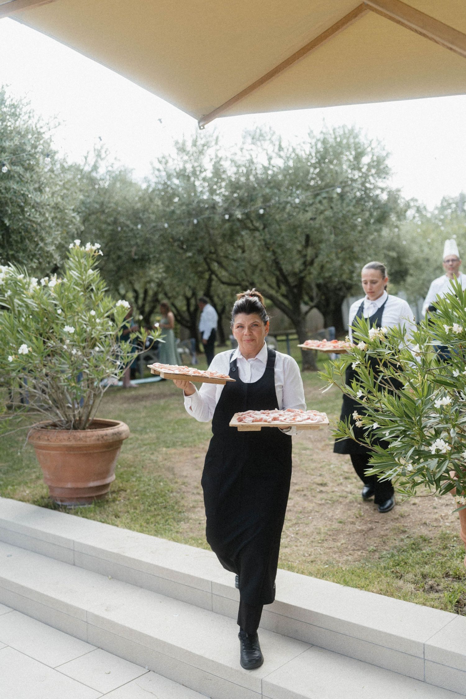 A waitress carries trays of food through a garden at an outdoor catering event.