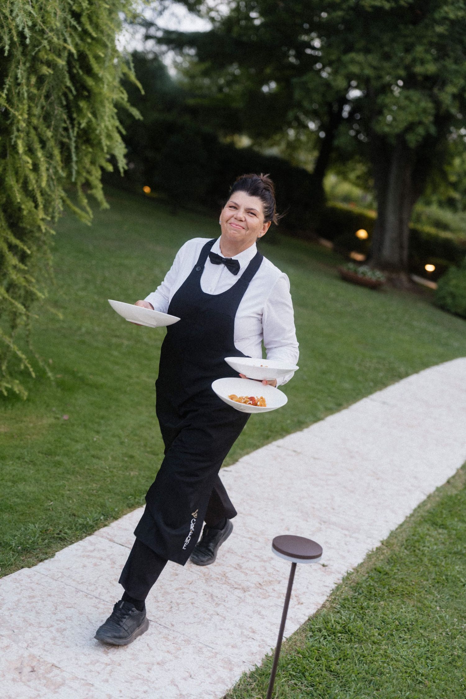 A smiling waitress in black apron and bow tie carries two plates along a garden path at an outdoor event.