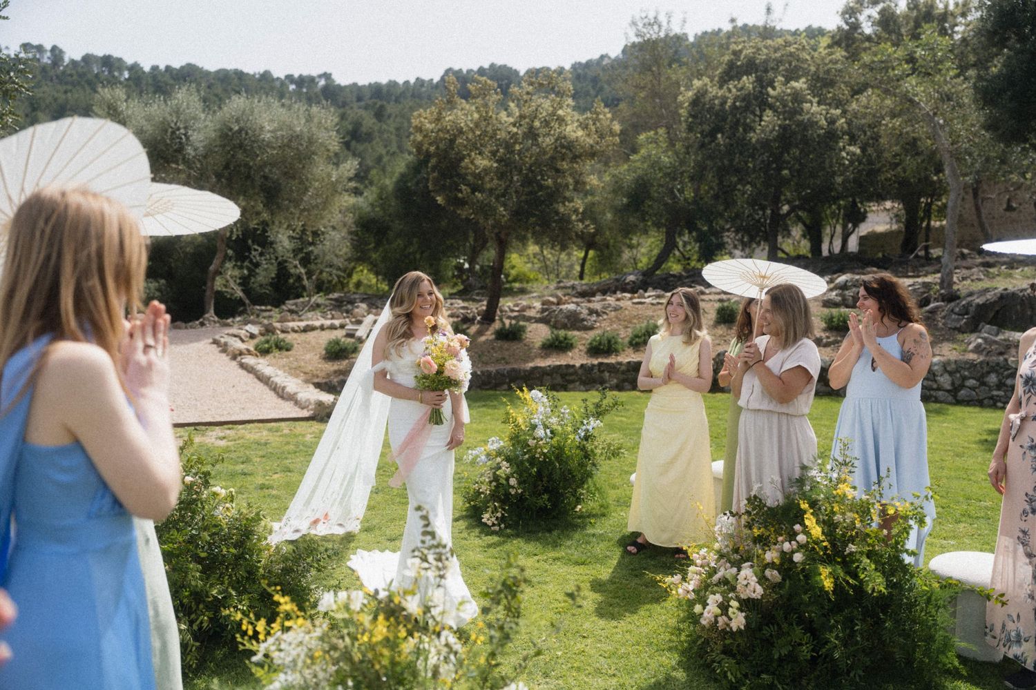 Bride in white gown with veil holds bouquet while bridesmaids in pastel dresses hold parasols at outdoor wedding.