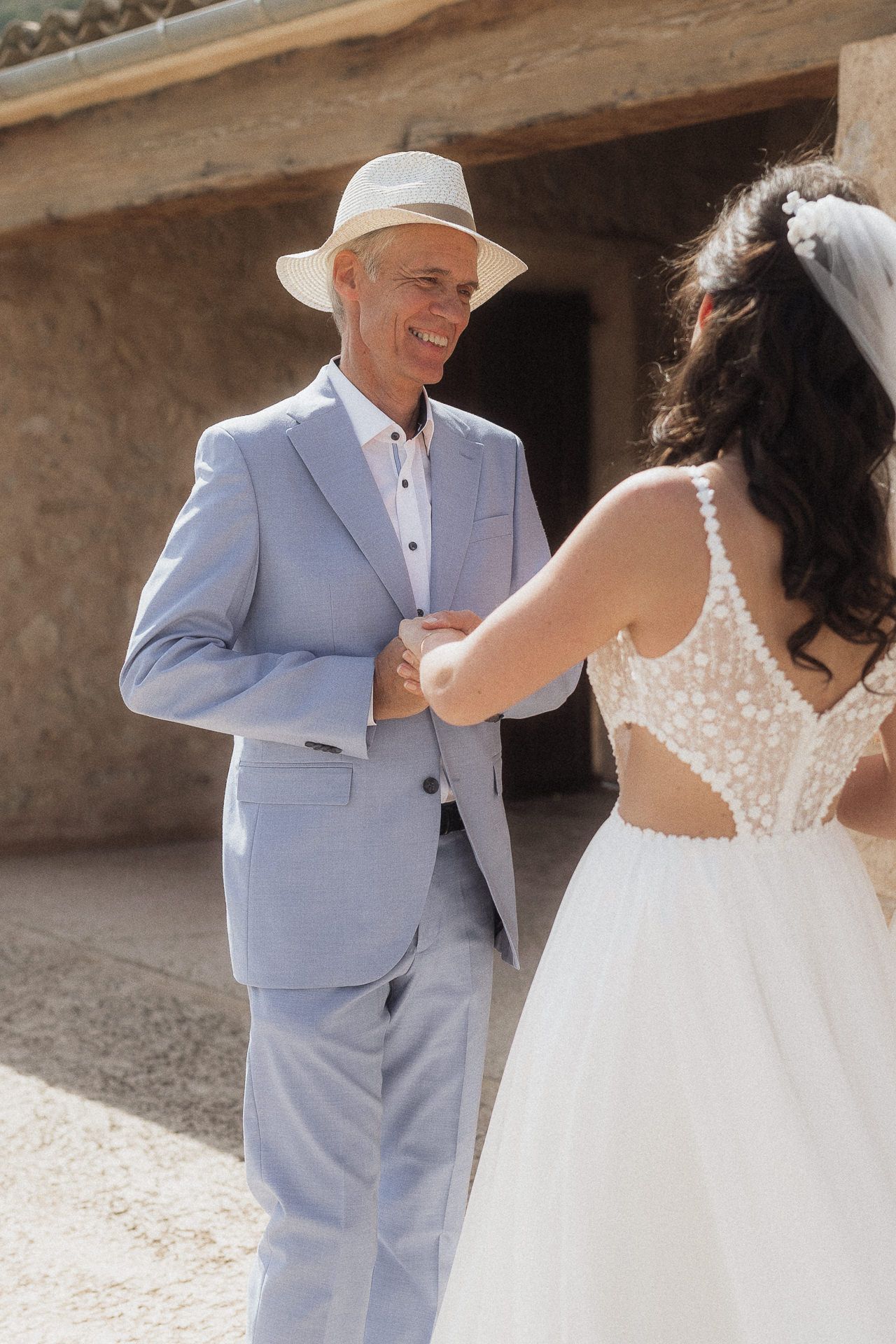 A smiling groom in a light blue suit and white hat faces a bride in a lace wedding dress outdoors.