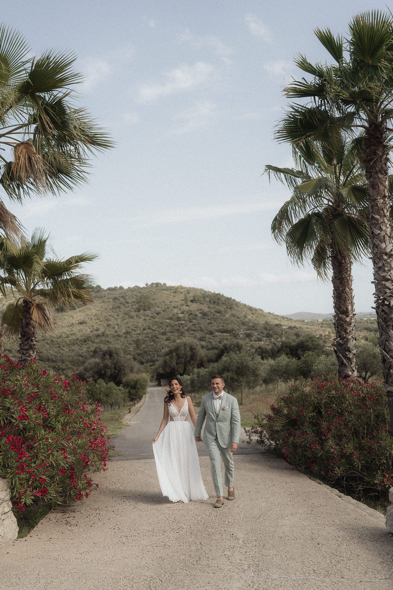 Bride and groom walk hand-in-hand down a palm-lined path with red flowers and rolling hills at an outdoor wedding.