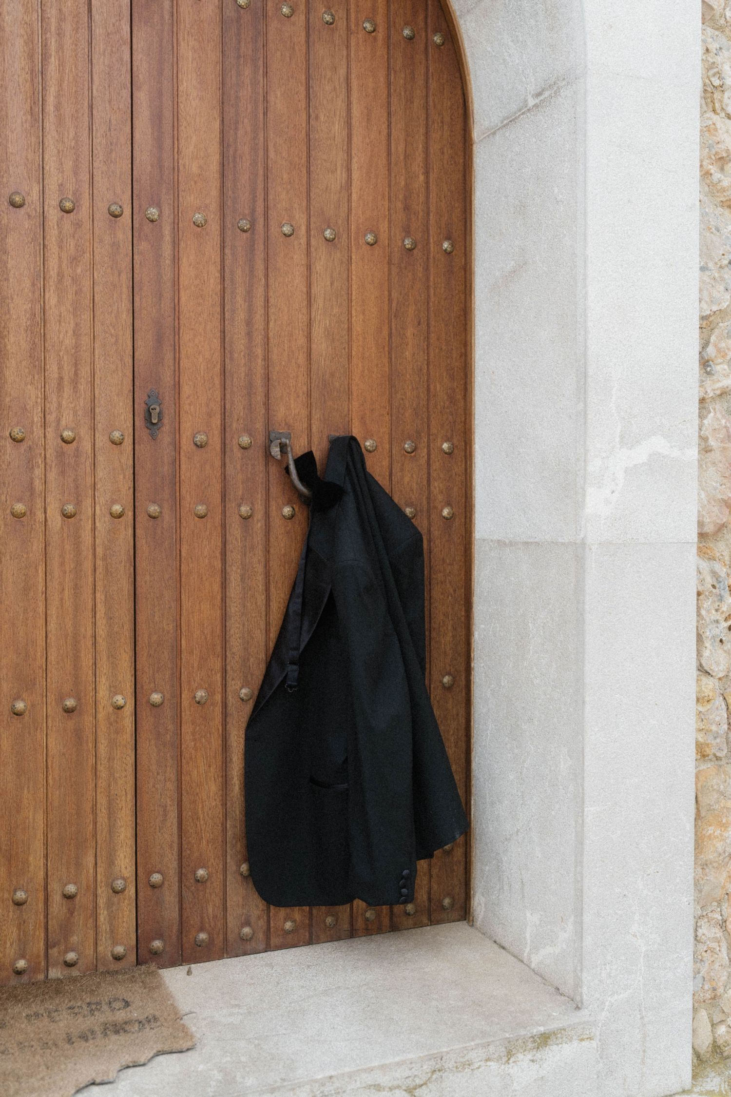 Black jacket hanging on ornate wooden arched door with metal studs on stone building.