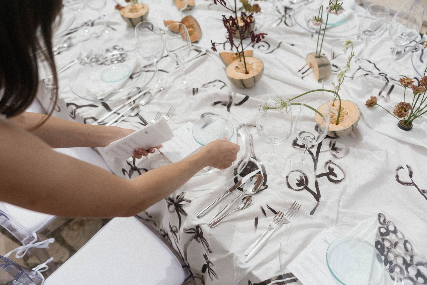 Woman setting an elegant table with white floral tablecloth, silverware, and small floral centerpieces.