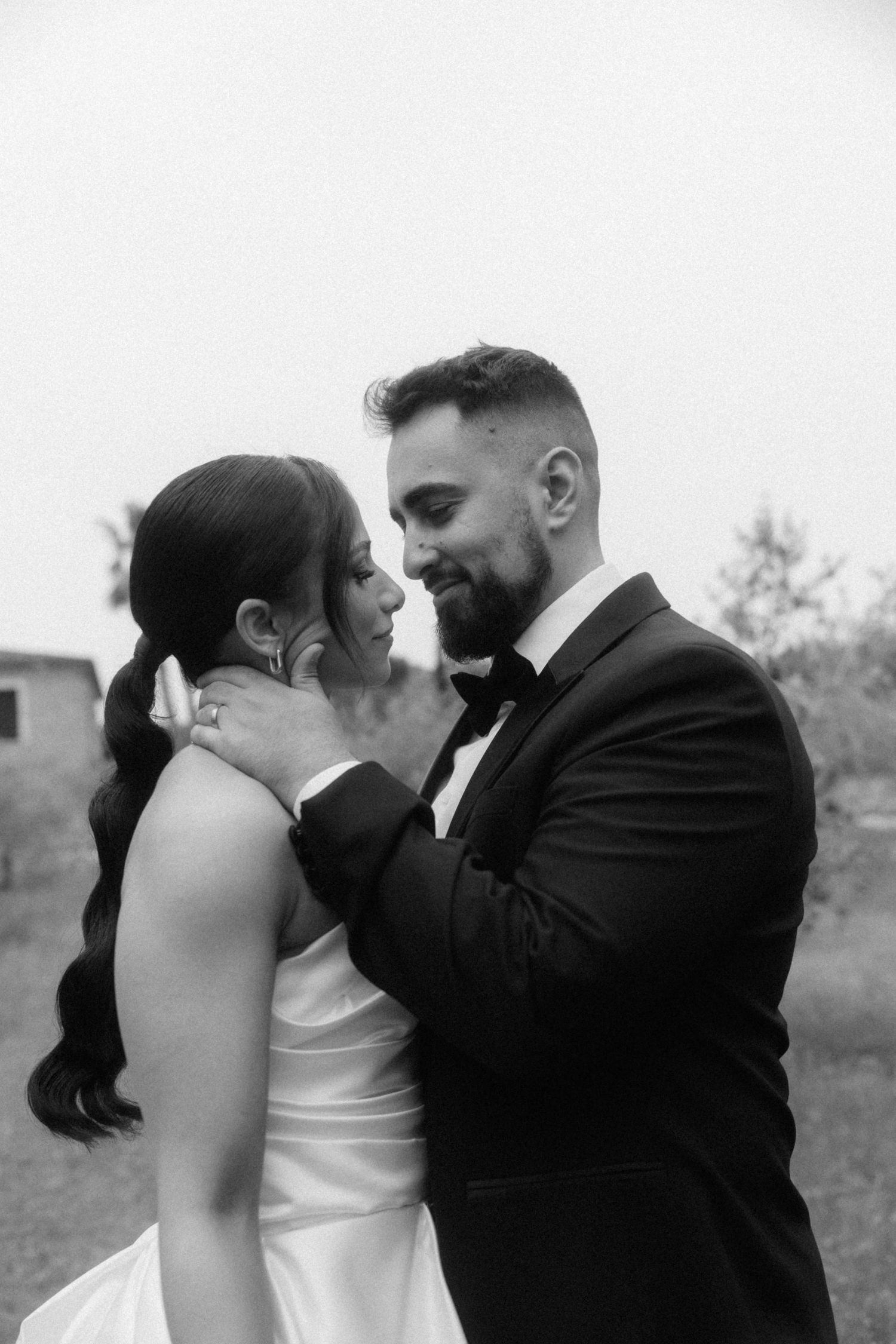 Black and white photo of smiling groom in tuxedo tenderly cupping brides face as they gaze at each other.