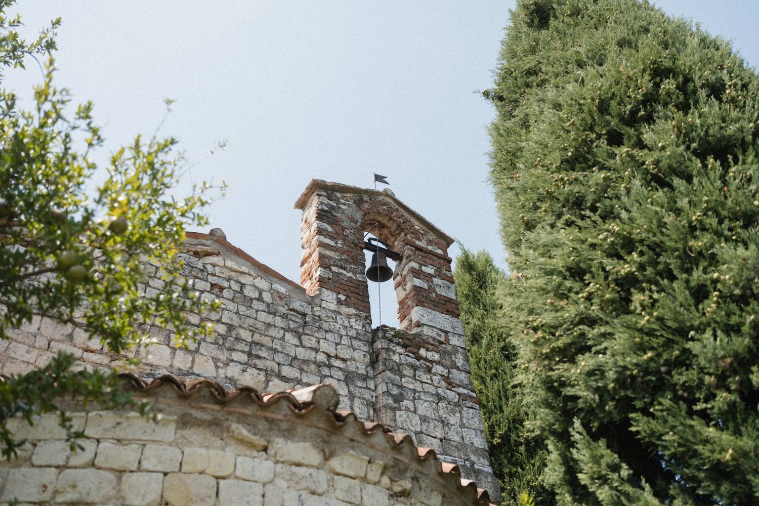 Stone chapel bell tower with cypress trees and lush greenery against a clear sky in a Mediterranean setting.