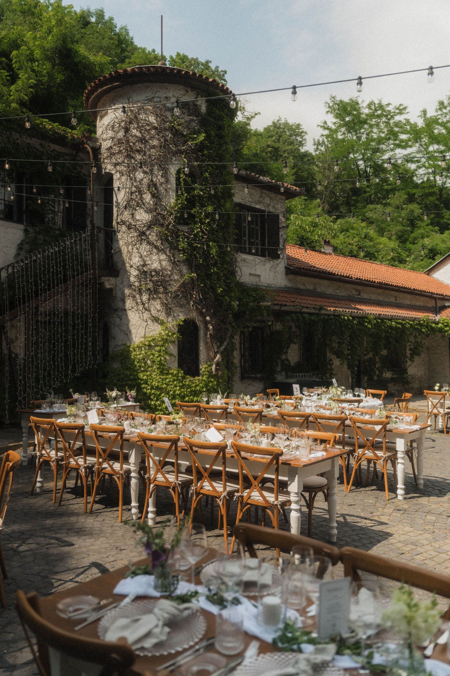 Elegant outdoor wedding reception with long tables, wooden chairs, and string lights at a rustic stone venue.