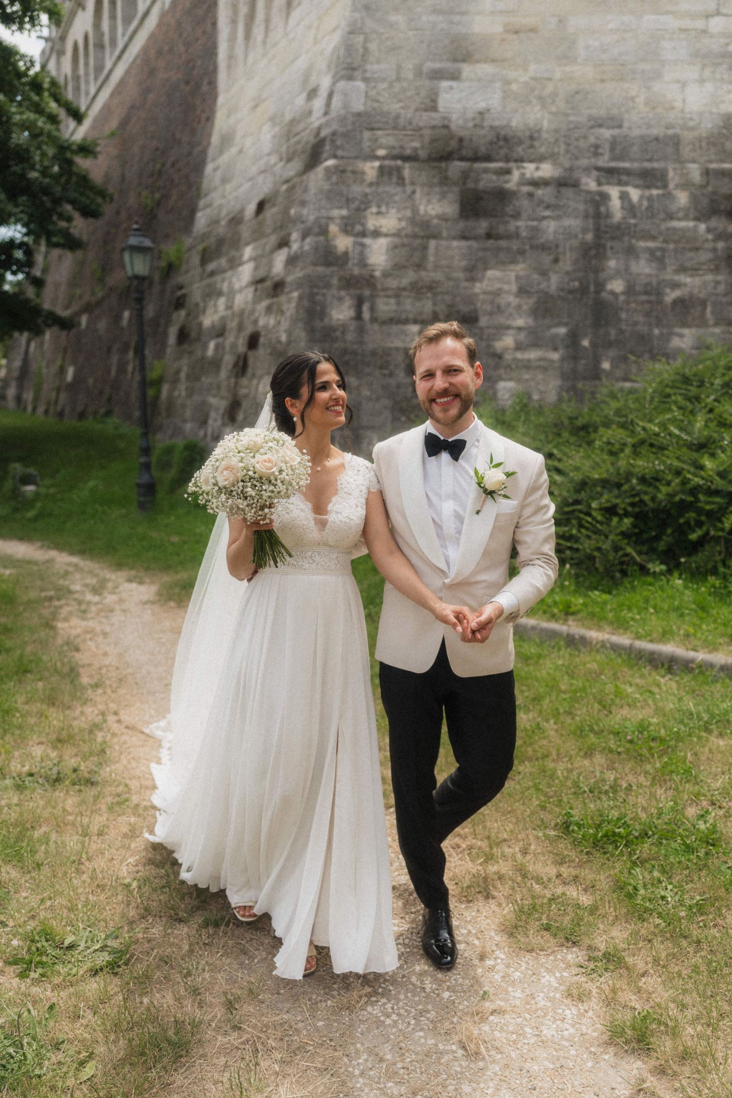 Happy newlywed couple strolling together near historic stone wall on wedding day.