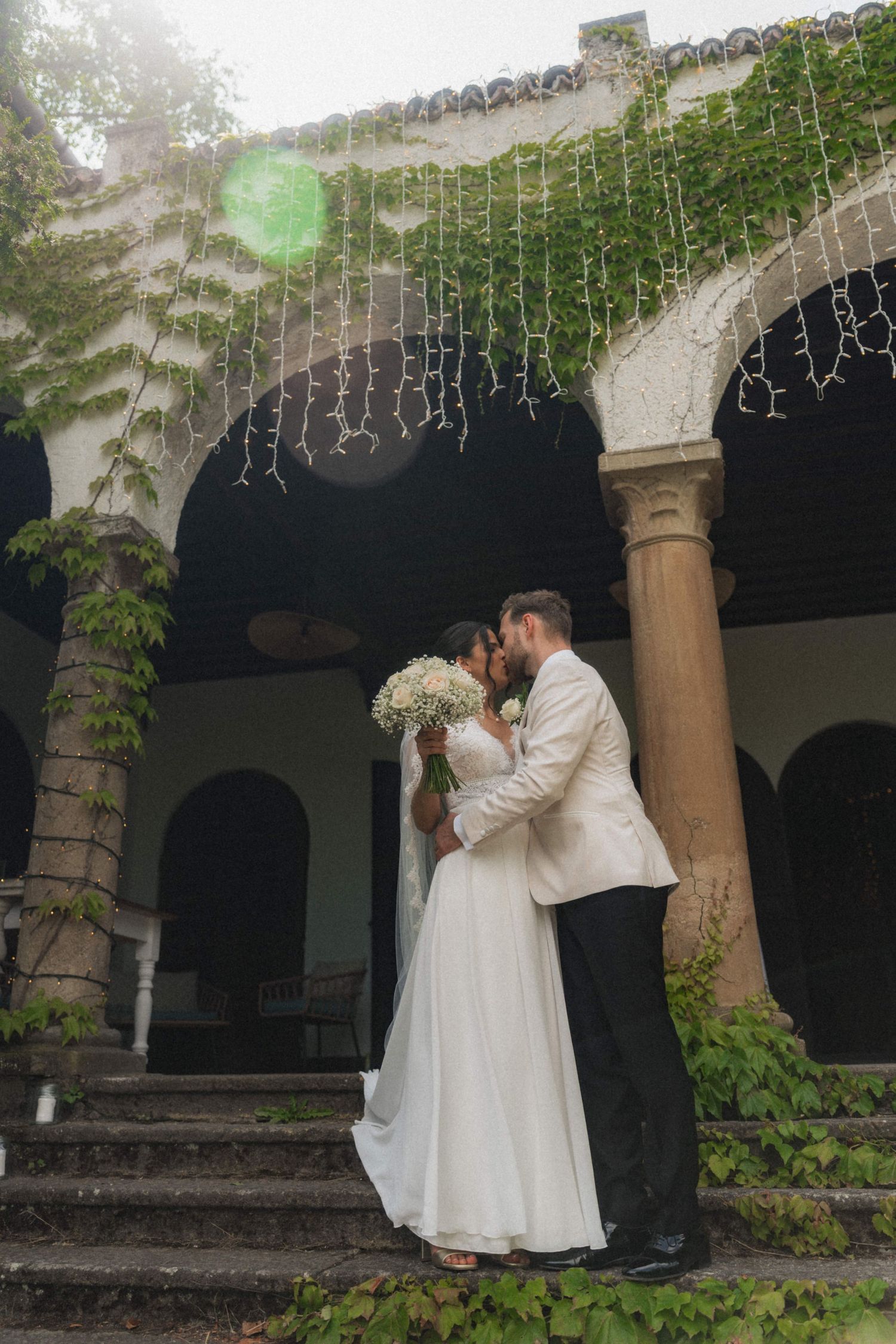 Newlyweds share a romantic kiss on stone steps of a vine-covered historic venue, bride holding white bouquet.