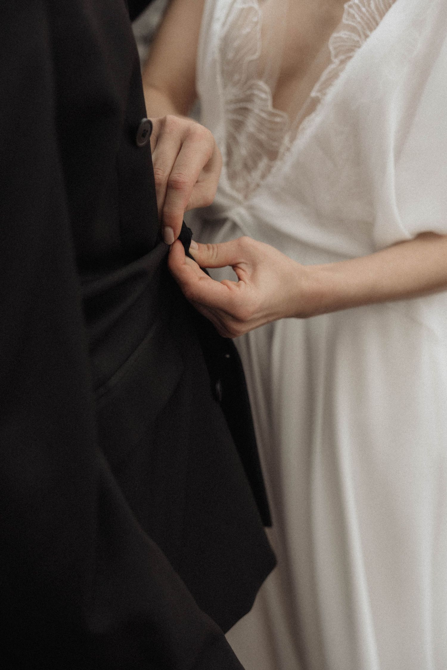 Close-up of bride in white lace dress holding grooms black suit jacket during wedding ceremony.
