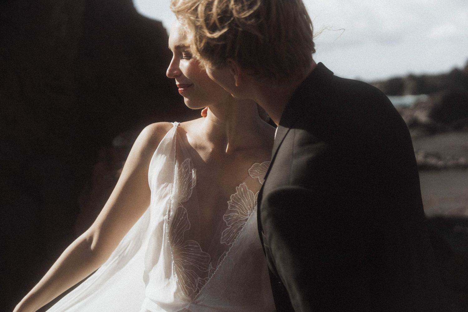 Bride in lace wedding dress leans against groom in black suit at rocky coastal setting.