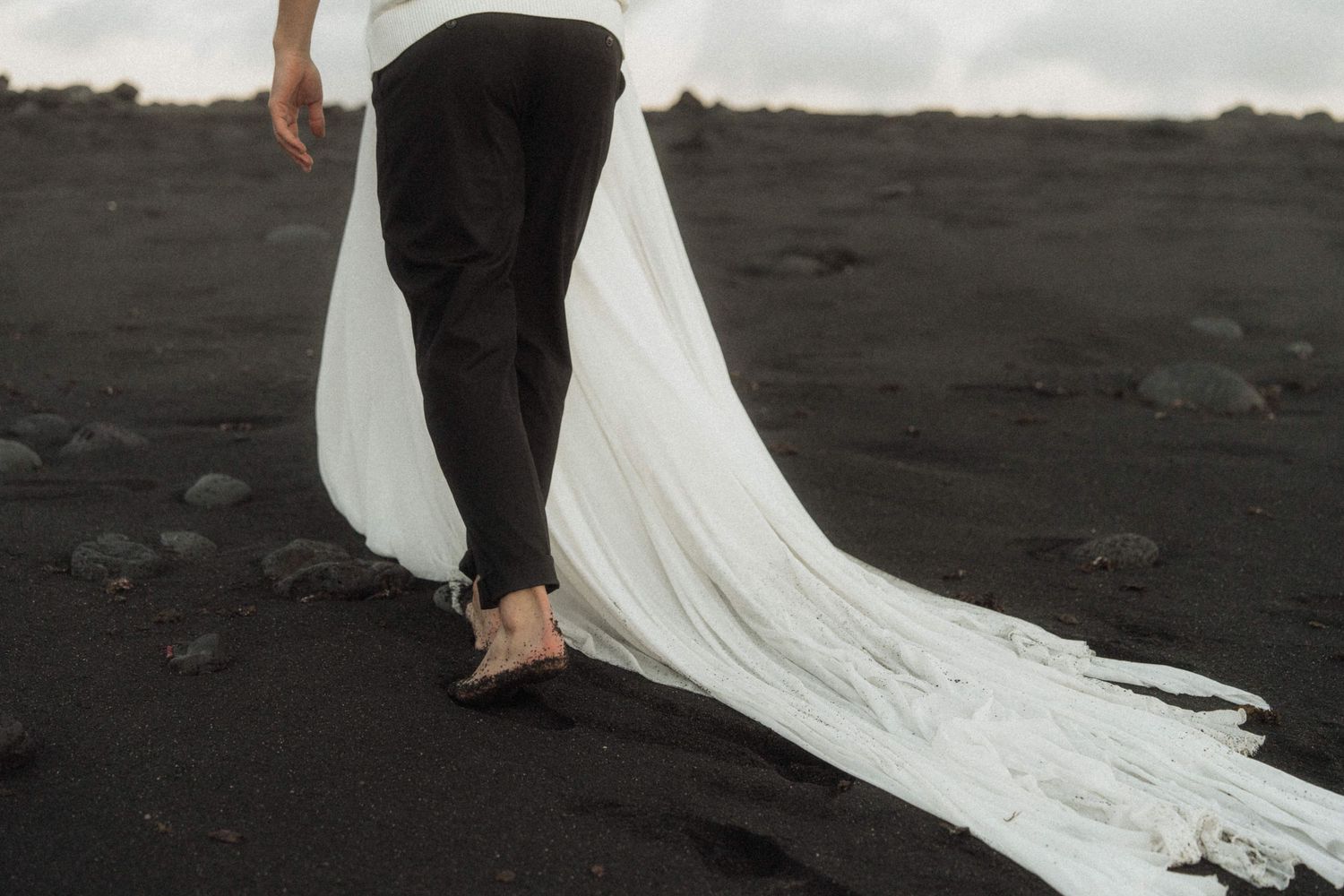 Bride walking barefoot on black sand beach with flowing white wedding dress train.
