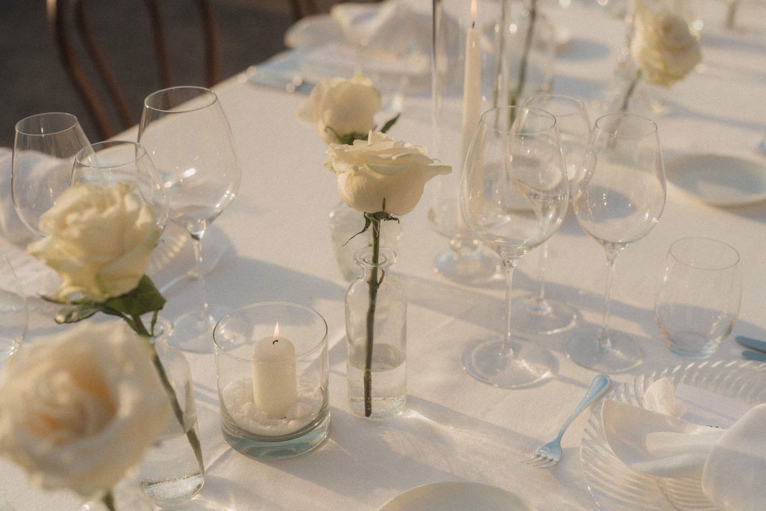 Elegant white wedding table setting with ivory roses, candles, and crystal glassware on a white linen tablecloth.