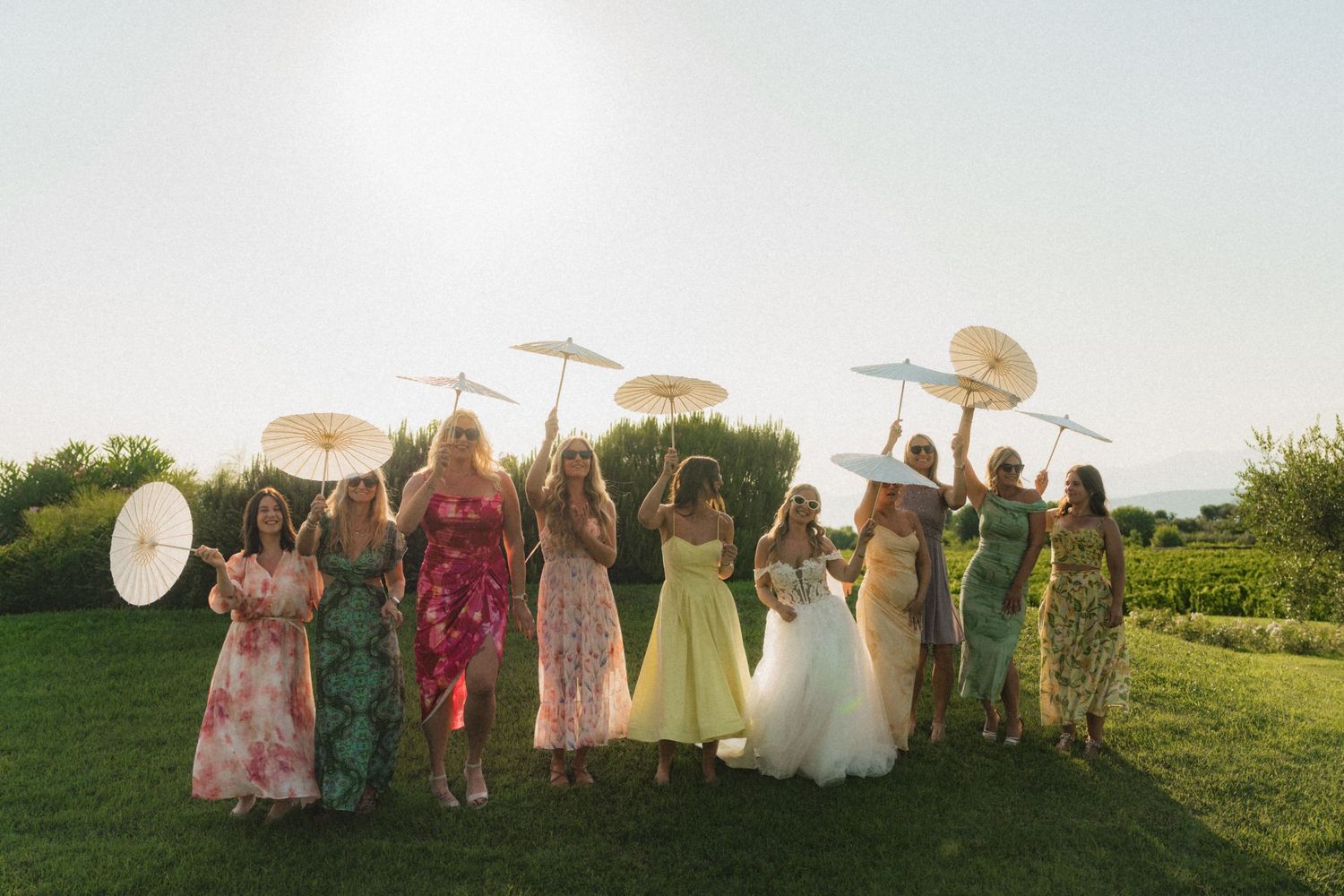 Bridal party in colorful floral dresses raising white parasols together in sunny garden.