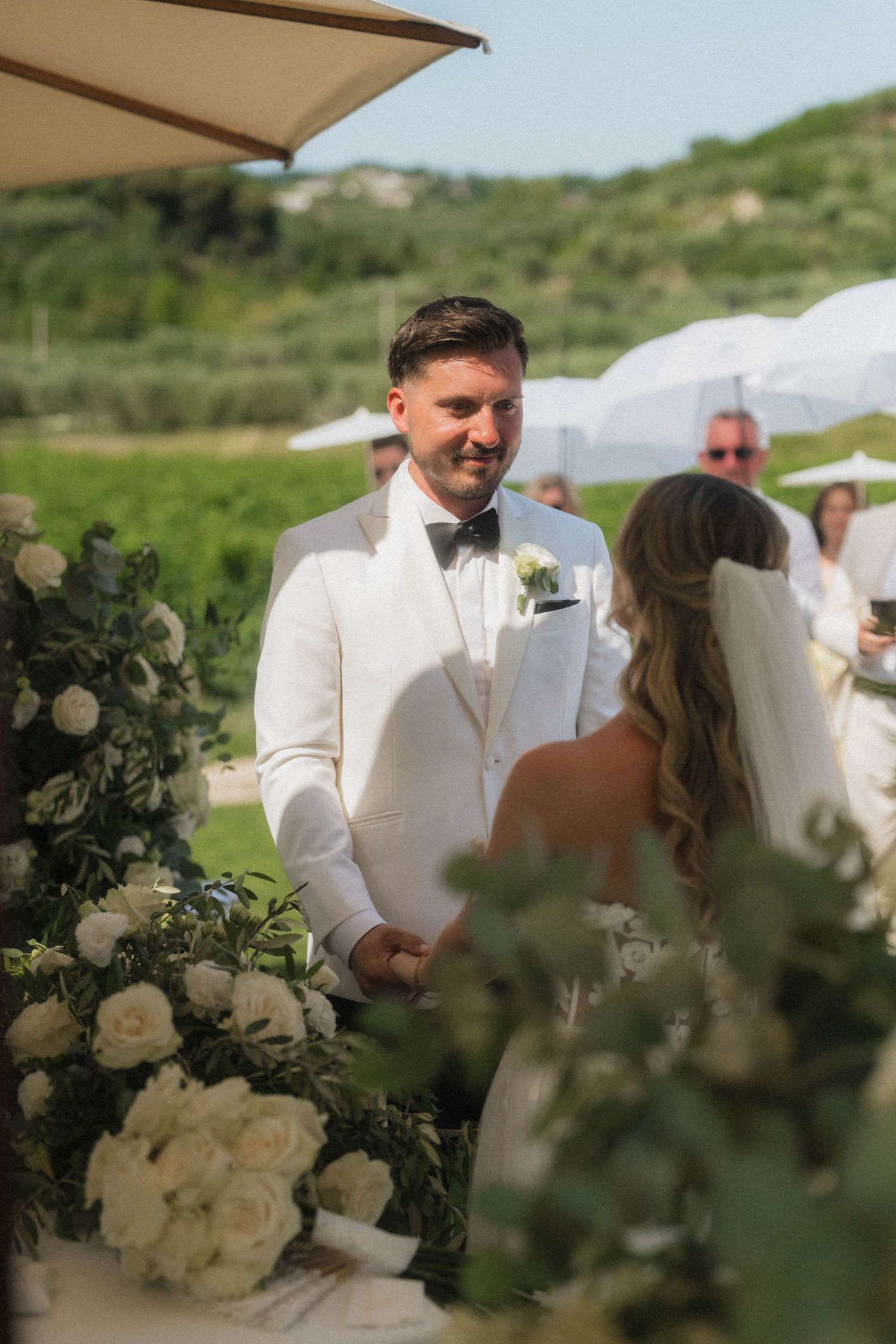 Groom in white tuxedo smiling at bride during outdoor wedding ceremony with white floral arrangements.