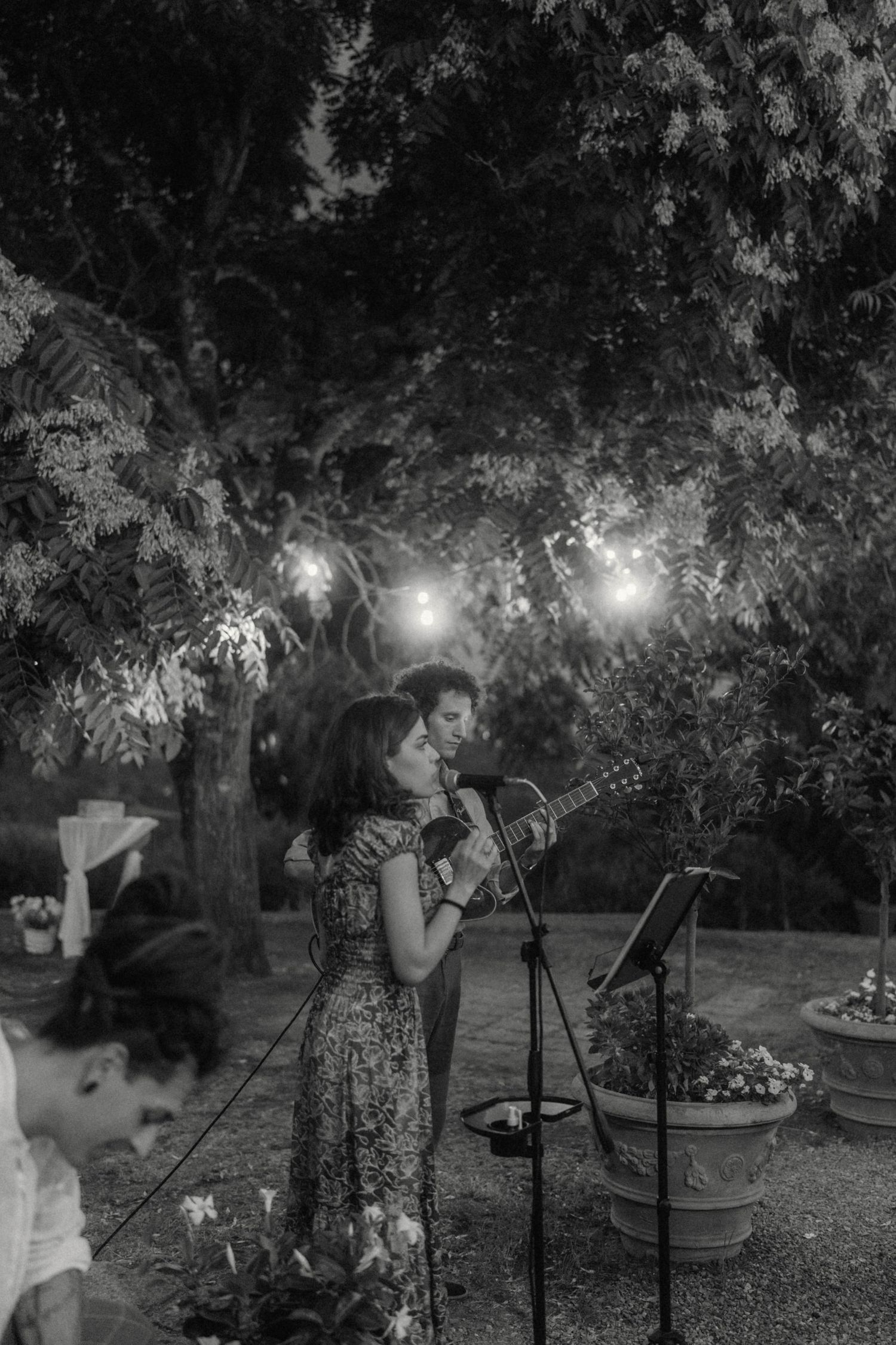 Black and white photo of a female vocalist and guitarist performing outdoors under string lights at a wedding reception.