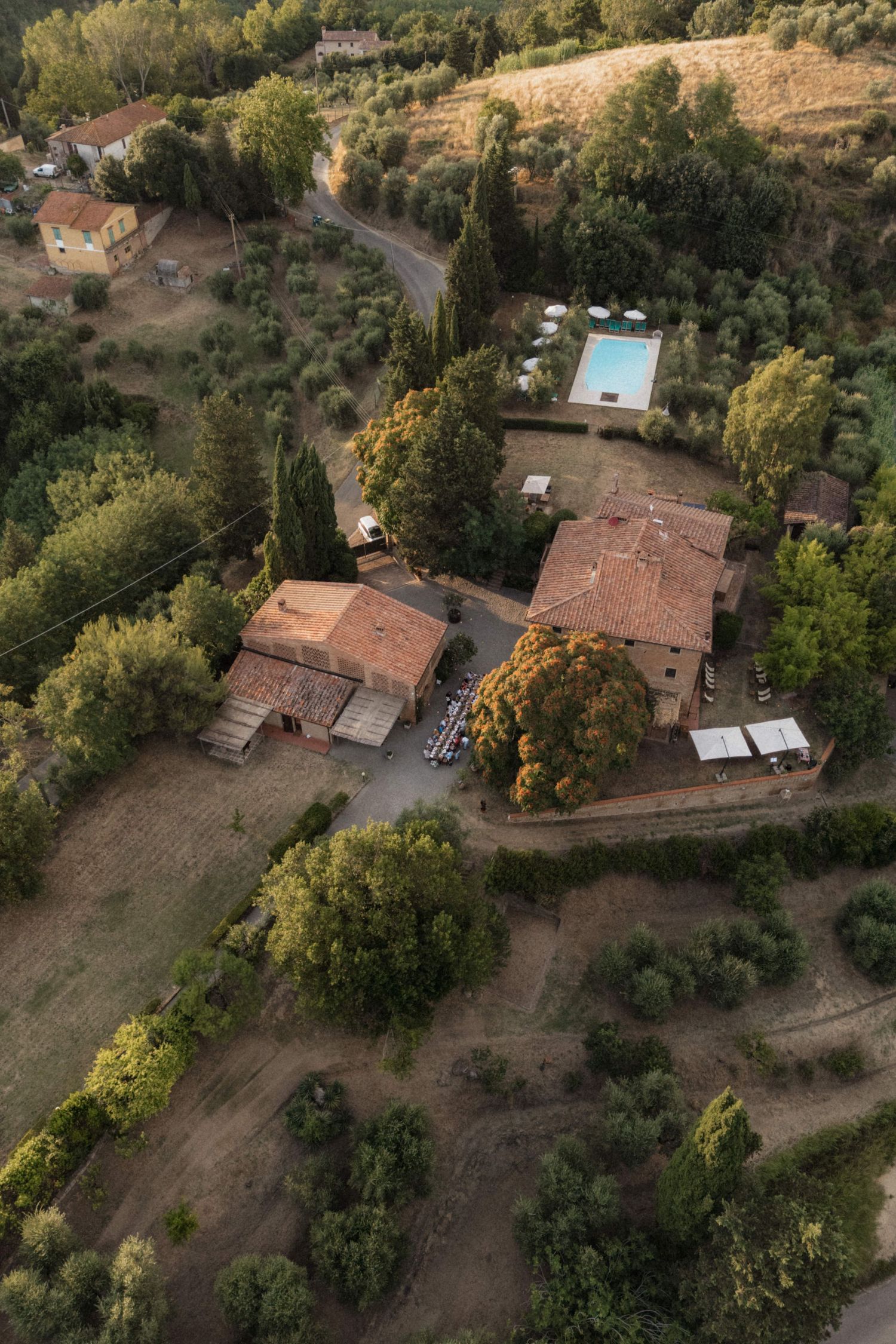 Drone aerial view of a rustic Tuscan farmhouse estate with terracotta roofs, pool, and lush surrounding greenery.