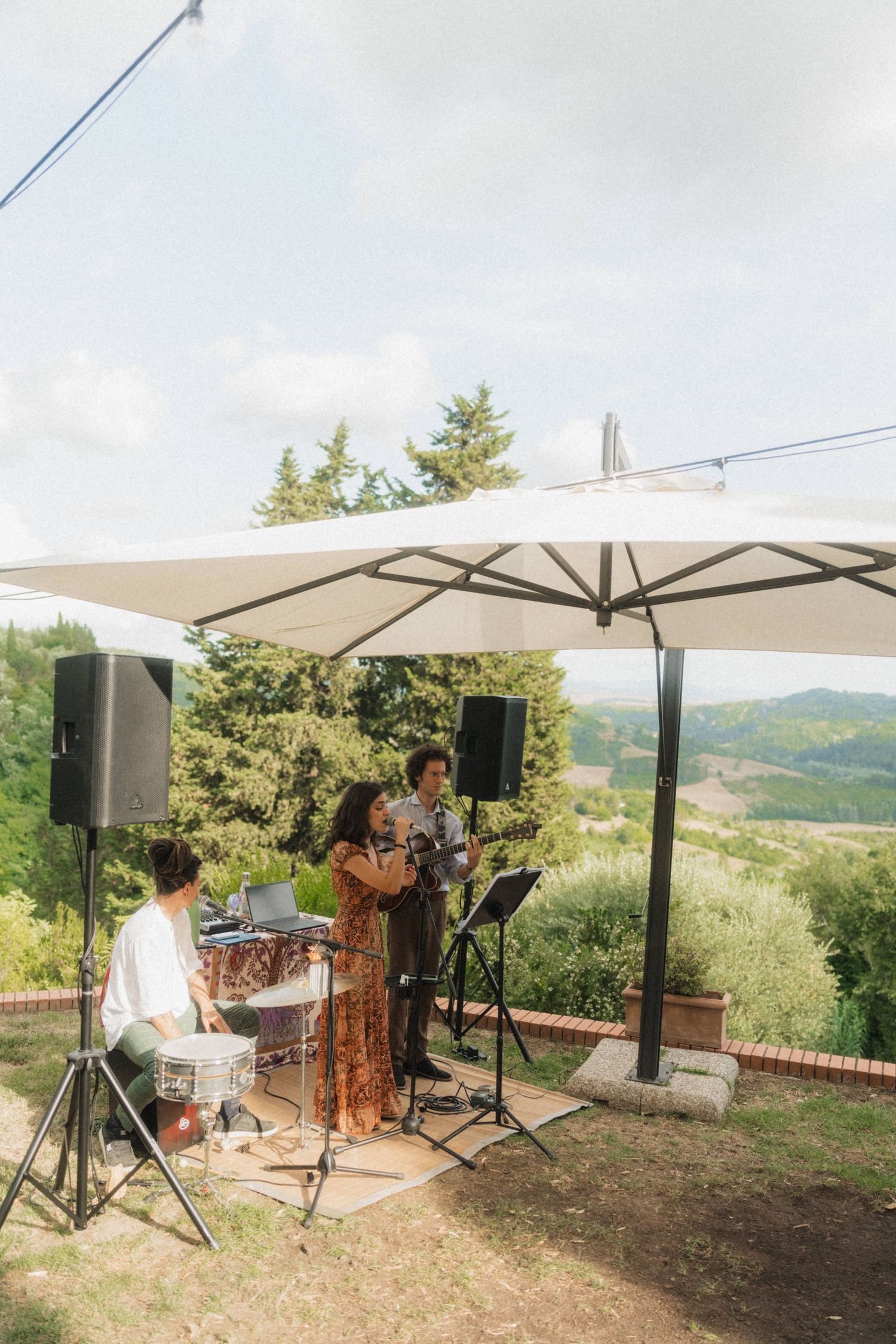 Live band performing under white umbrella at outdoor Tuscan wedding with scenic countryside view.