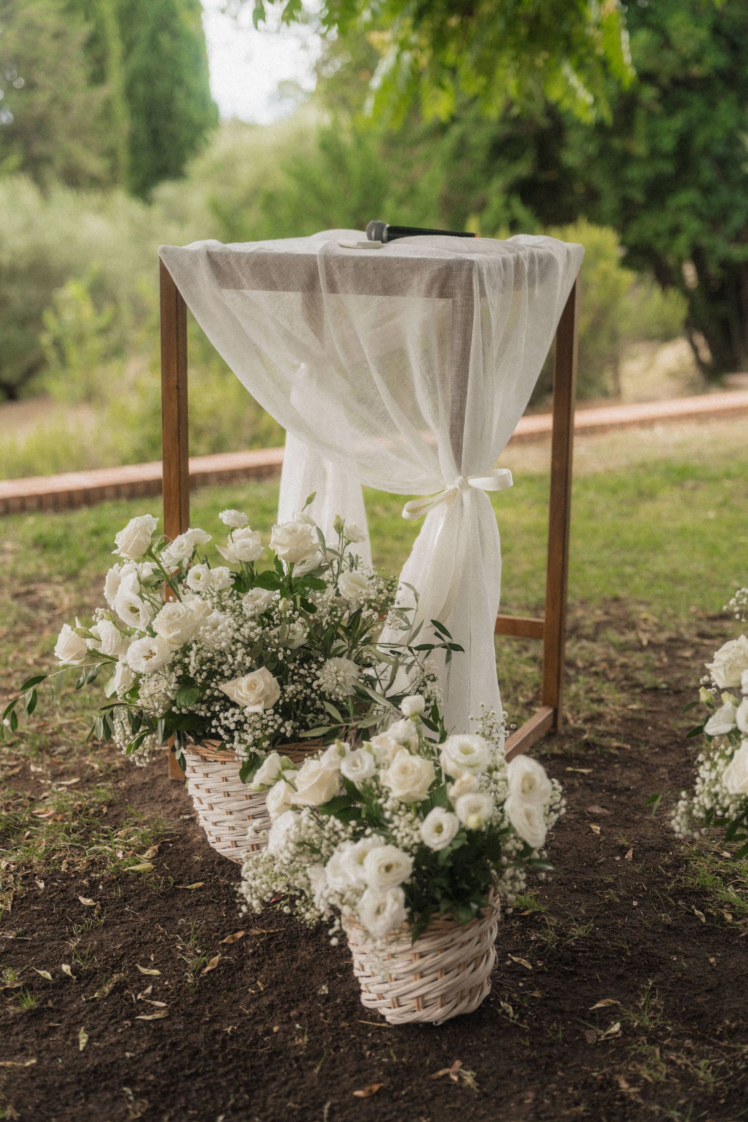 Rustic wooden wedding altar draped with white sheer fabric and adorned with white rose floral arrangements in wicker baskets.