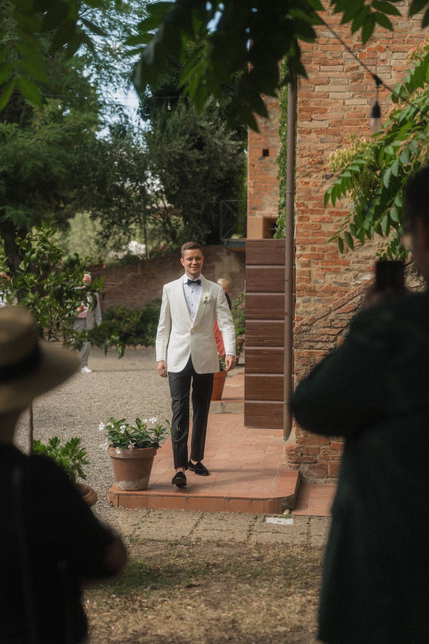 Groom in white tuxedo jacket walks toward guests at an outdoor rustic brick venue wedding.
